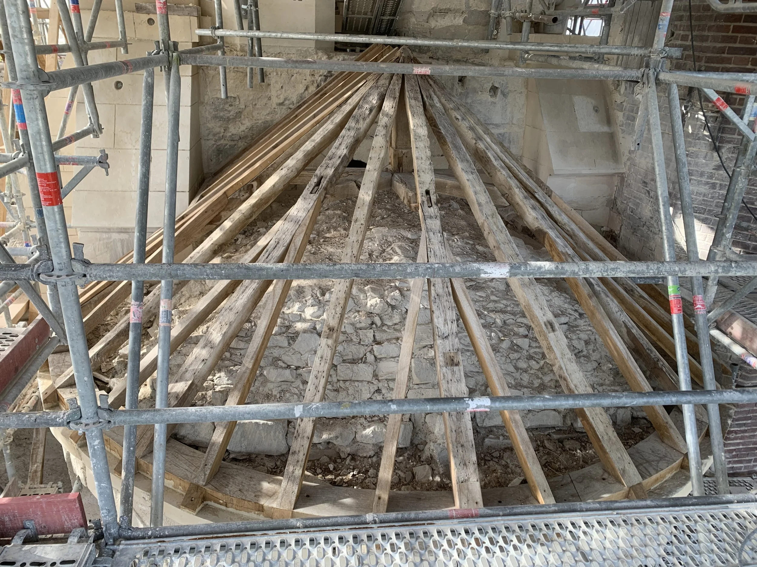 Construction en cours avec un échafaudage en métal autour d'une structure en bois et des pierres, probablement dans un bâtiment ancien en restauration.