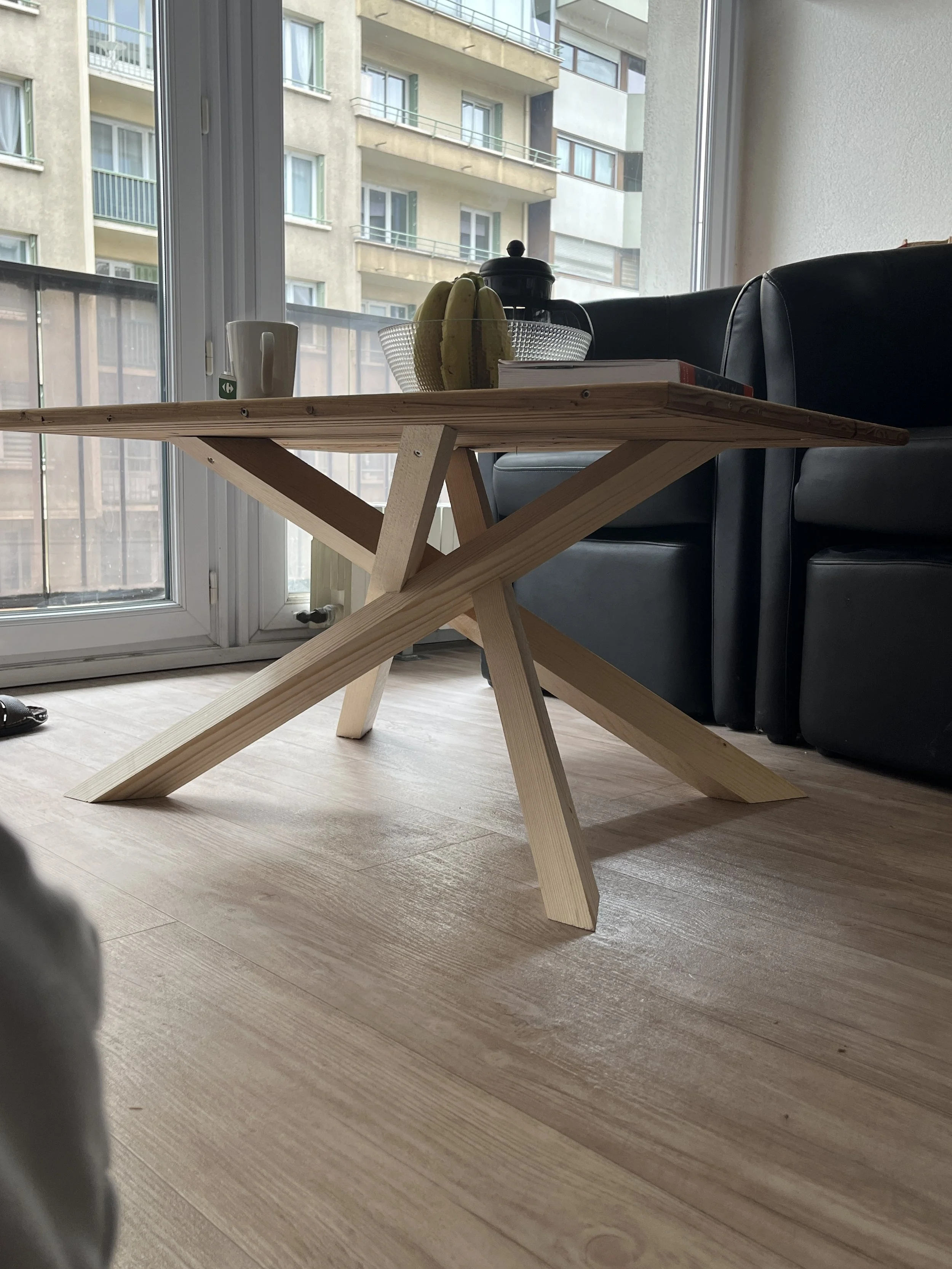 Table en bois clair dans un appartement avec vue sur un bâtiment résidentiel, une tasse blanche, une armoire à café, une corbeille avec des bananes, et des livres. Sol en bois clair.