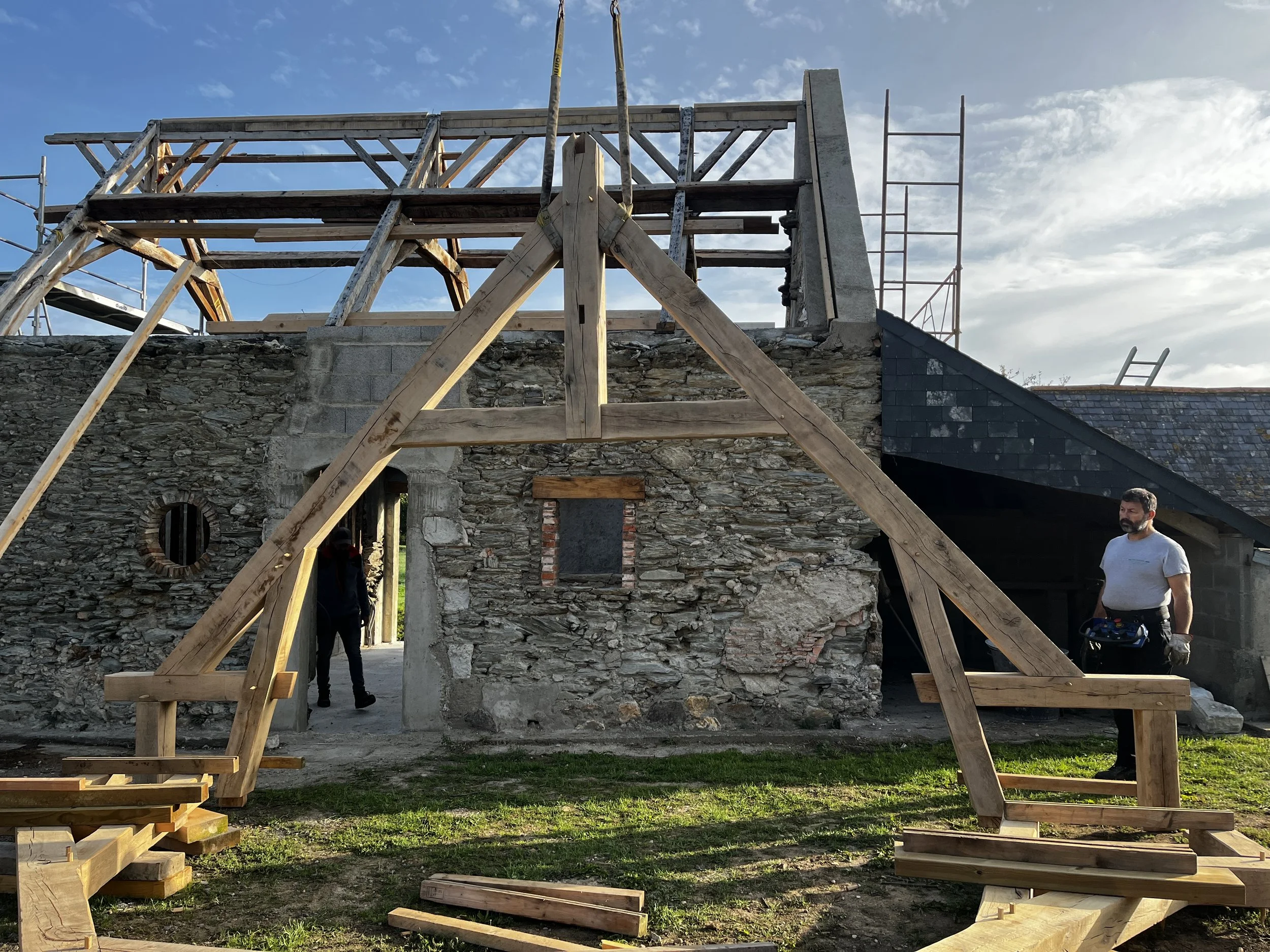 Une structure en bois en cours de construction contre un mur en pierre, avec un ciel partiellement nuageux en arrière-plan et deux hommes présents.