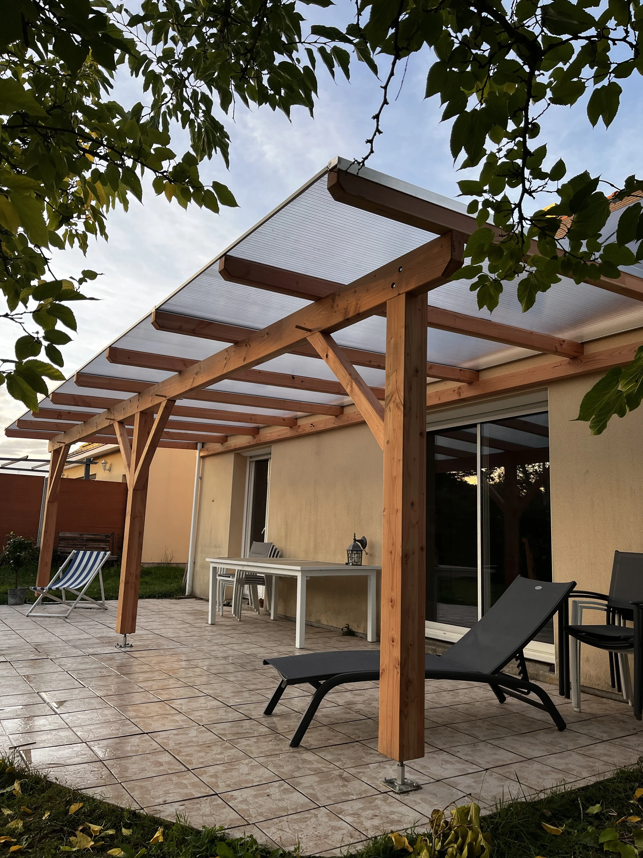 Une terrasse extérieure avec un auvent en bois, des chaises pliantes, une table blanche, un lit solaire noir, et des plantes vertes, sous un ciel partiellement nuageux.