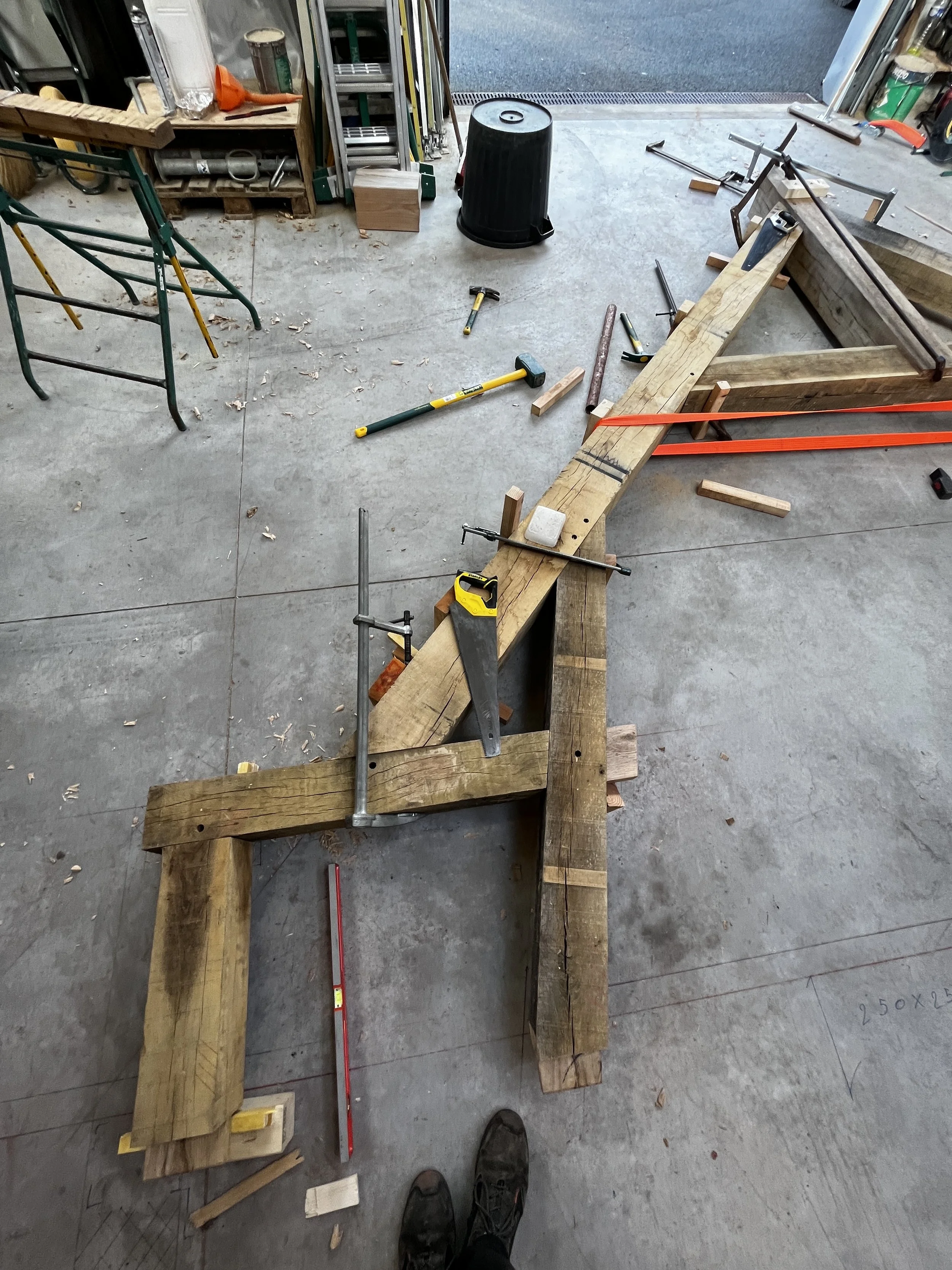 Intérieur d'un atelier de menuiserie avec des outils en bois, une échelle, une poubelle noire, un marteau, un niveau, et un plan de travail en bois en cours de construction.