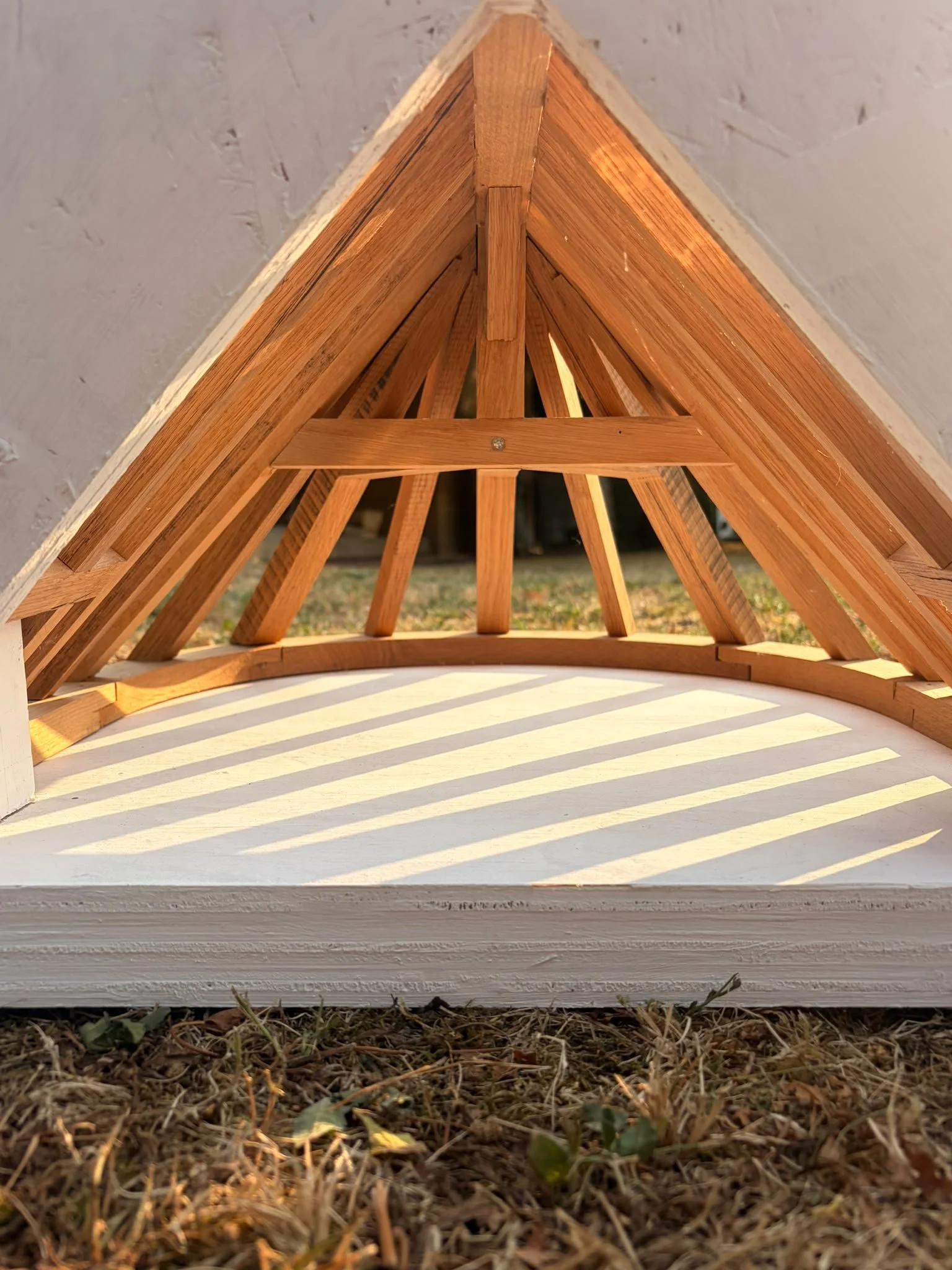 Une structure en bois formant un toit triangulaire située à l'extérieur, avec de l'ombre créée par la lumière du soleil, et le sol recouvert de petites plantes et de terre.