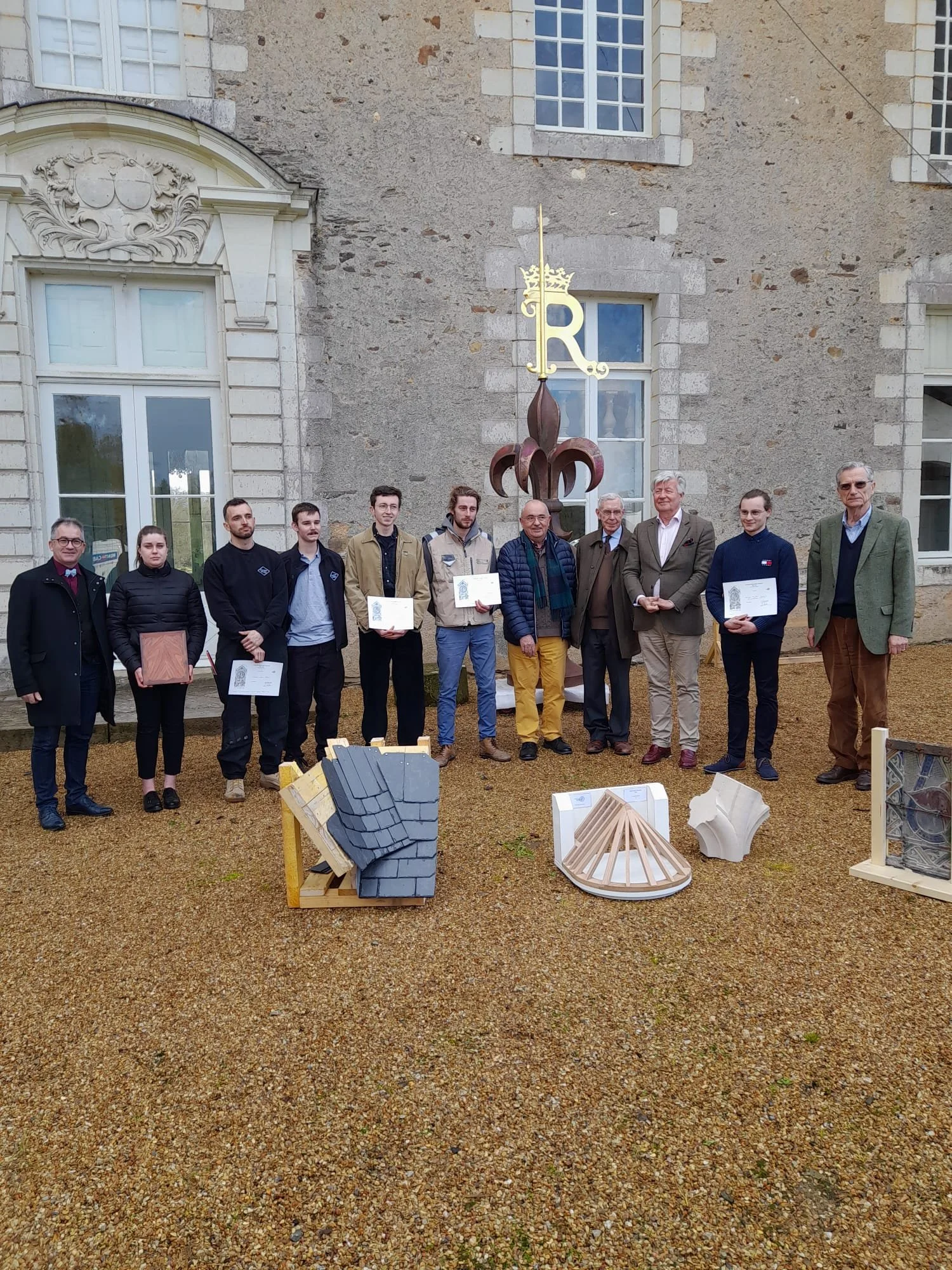 Groupe de personnes posant devant une sculpture de style artistique en extérieur, avec des maquettes de conception architecturale exposées sur le sol devant eux, devant un bâtiment en pierre avec plusieurs fenêtres.