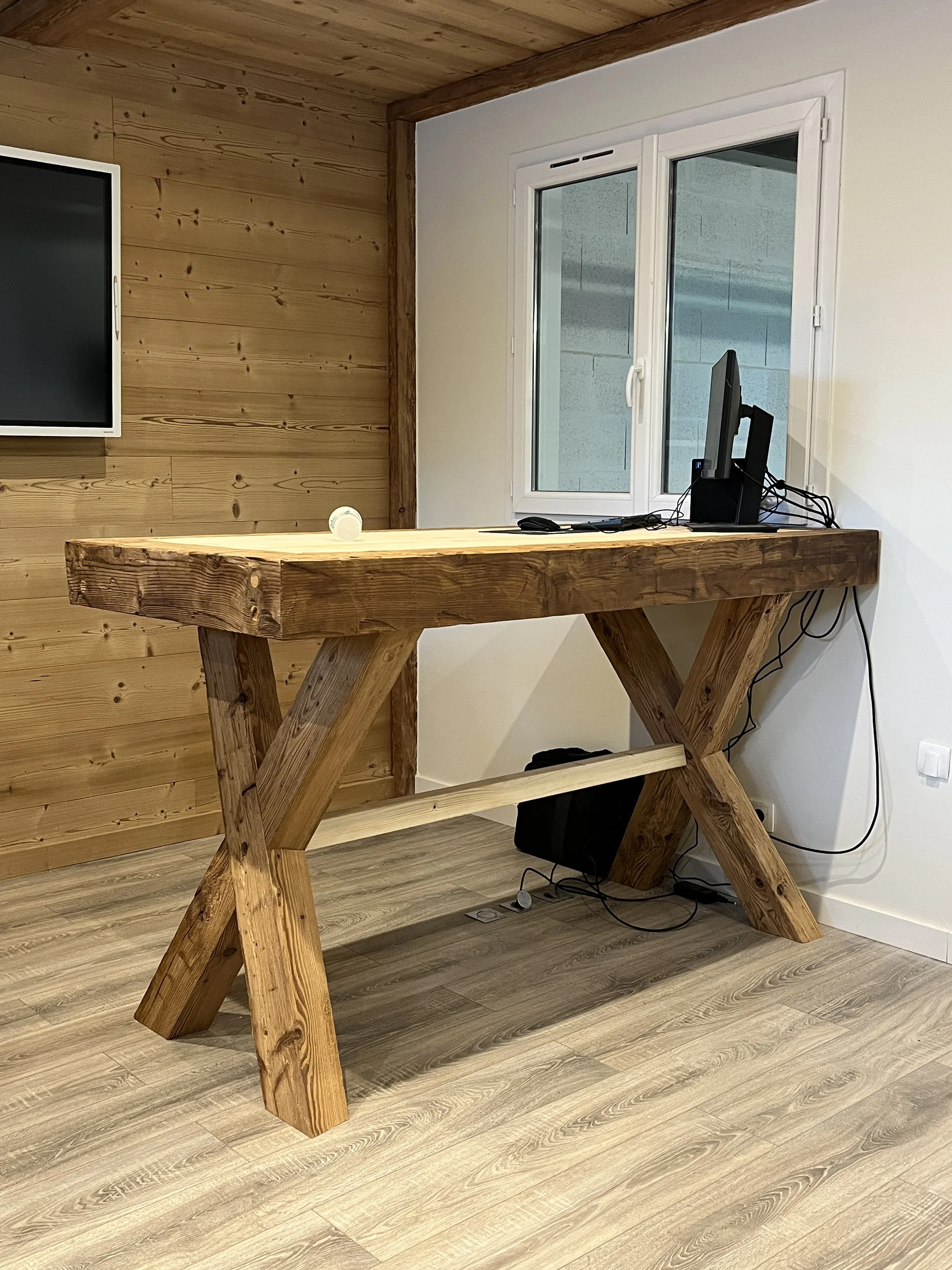 Un bureau en bois brut avec une dalle en bois massif, placé dans une pièce avec un mur en bois et un mur blanc. Sur le bureau, il y a un ordinateur avec un clavier et une souris, et un câble électrique enroulé. À côté du bureau, il y a une prise électrique au sol et un câble connecté à l'ordinateur.