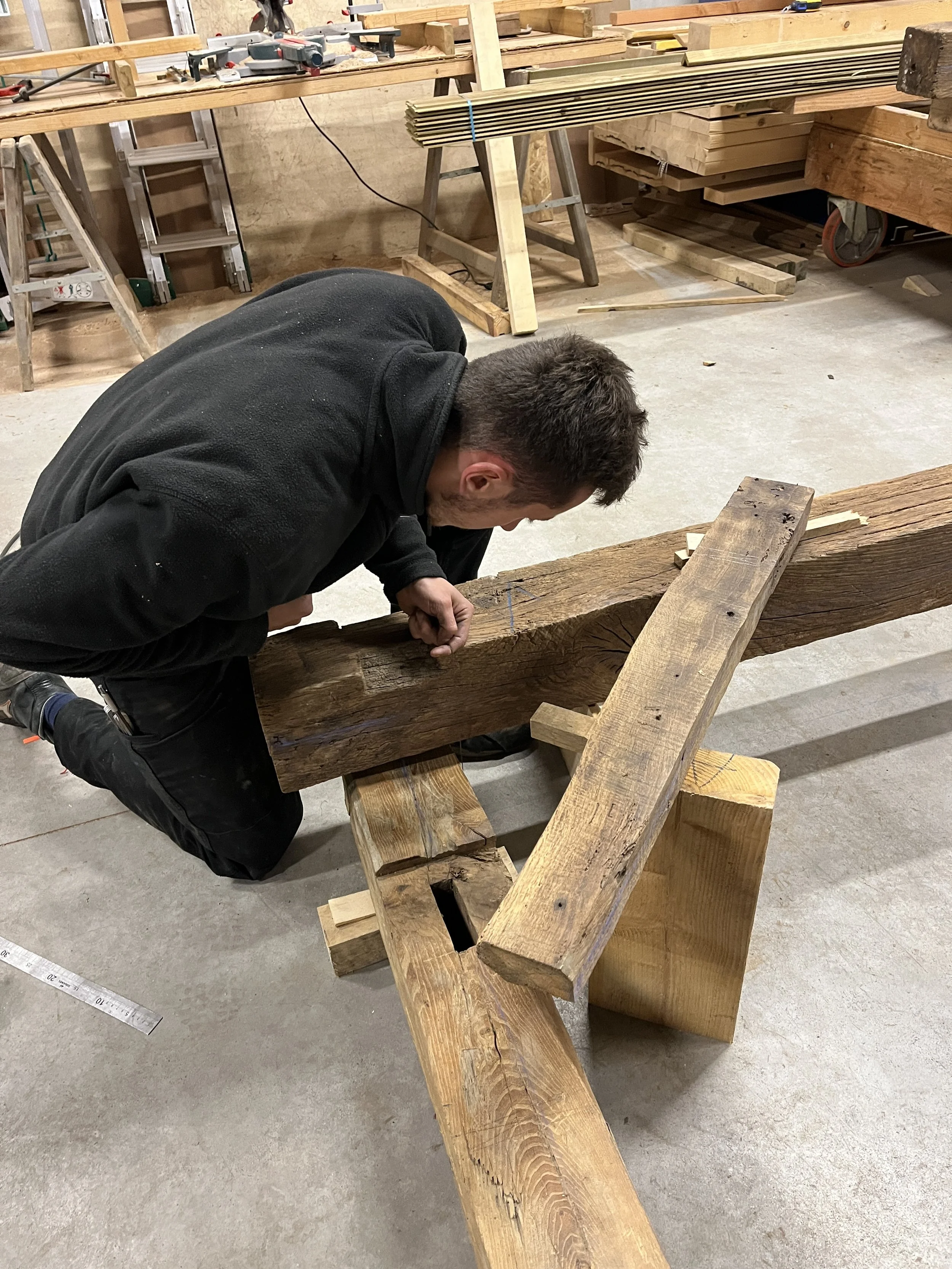 Un homme travaille sur une pièce de bois dans un atelier de menuiserie, entouré d'outils, de planches de bois et d'équipements de bricolage.