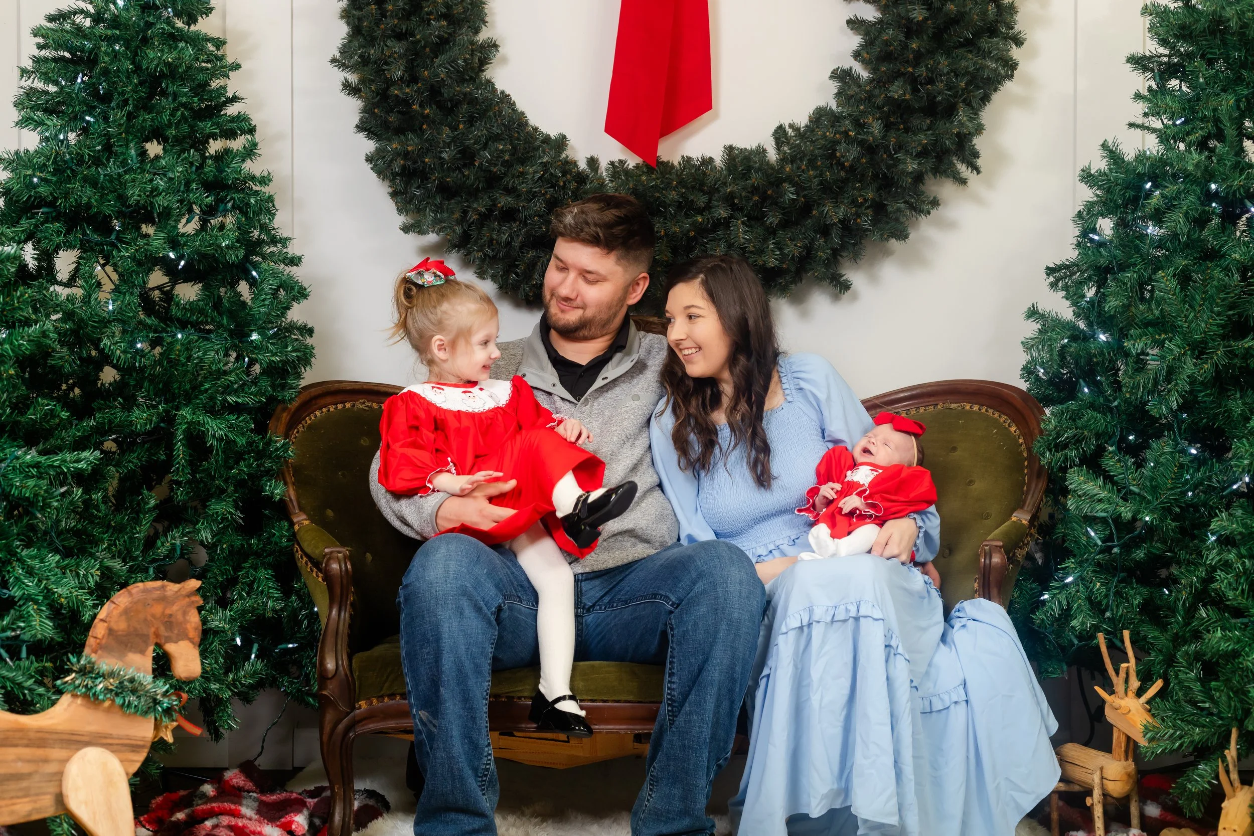 Family sitting on vintage sofa surrounded by Christmas trees, with a family of four including two young children dressed in holiday outfits, celebrating Christmas.