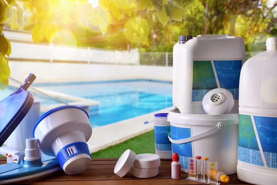 Pool cleaning chemicals and equipment on a wooden surface near a backyard swimming pool.