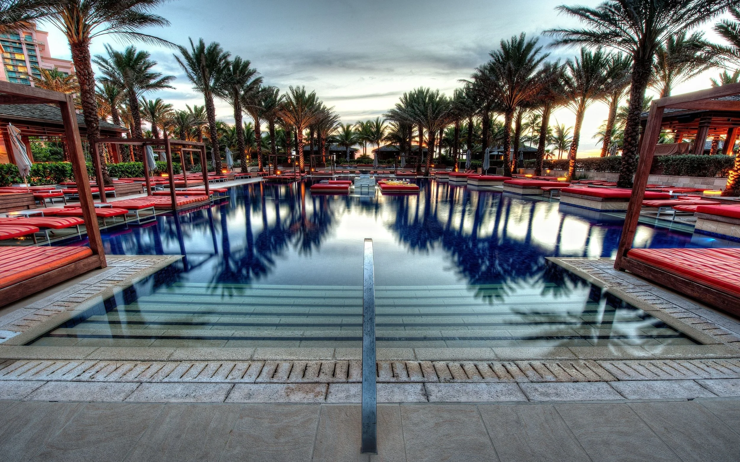 A resort swimming pool area at sunset, with palm trees lining the pool and red lounge chairs with umbrellas around it.