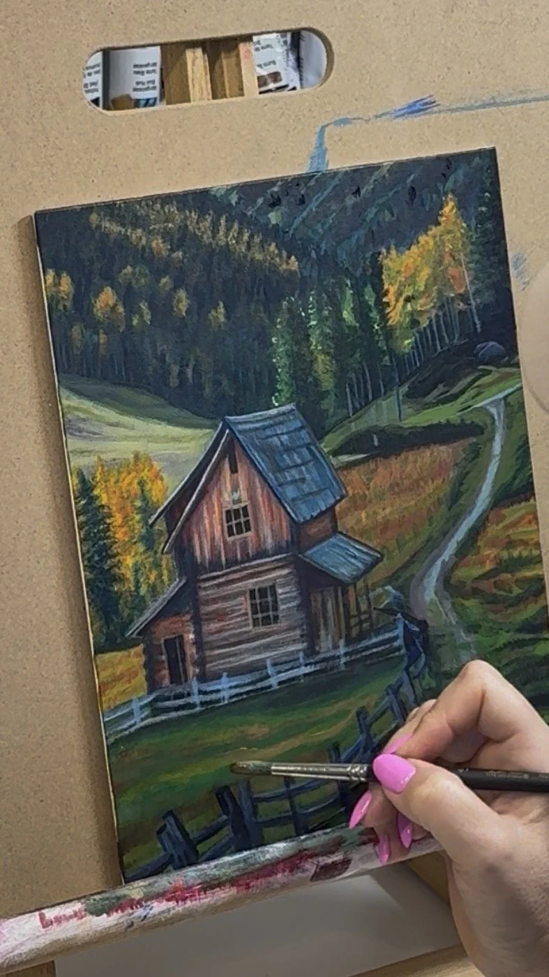A person painting a landscape of a countryside scene featuring a rustic wooden barn, a winding path, green fields, and colorful trees during autumn, on a canvas.