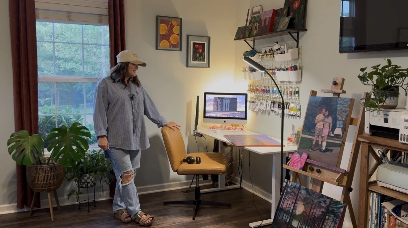 Woman standing next to a window in a craft room, with artwork and a computer on a desk, and painting supplies and plants around the room.