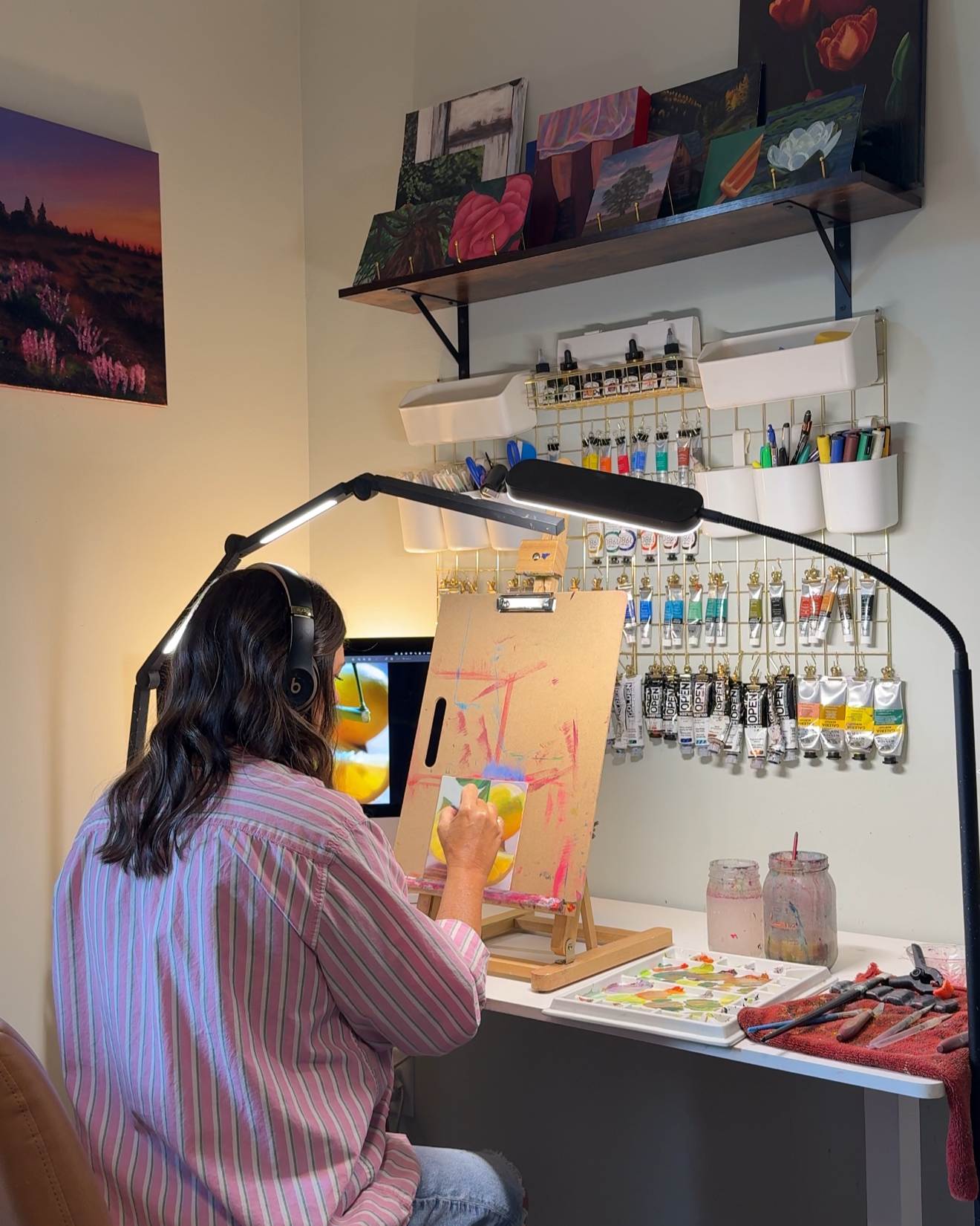 Woman painting on a canvas in an art studio with art supplies, paintings, and jars of paint on the desk.