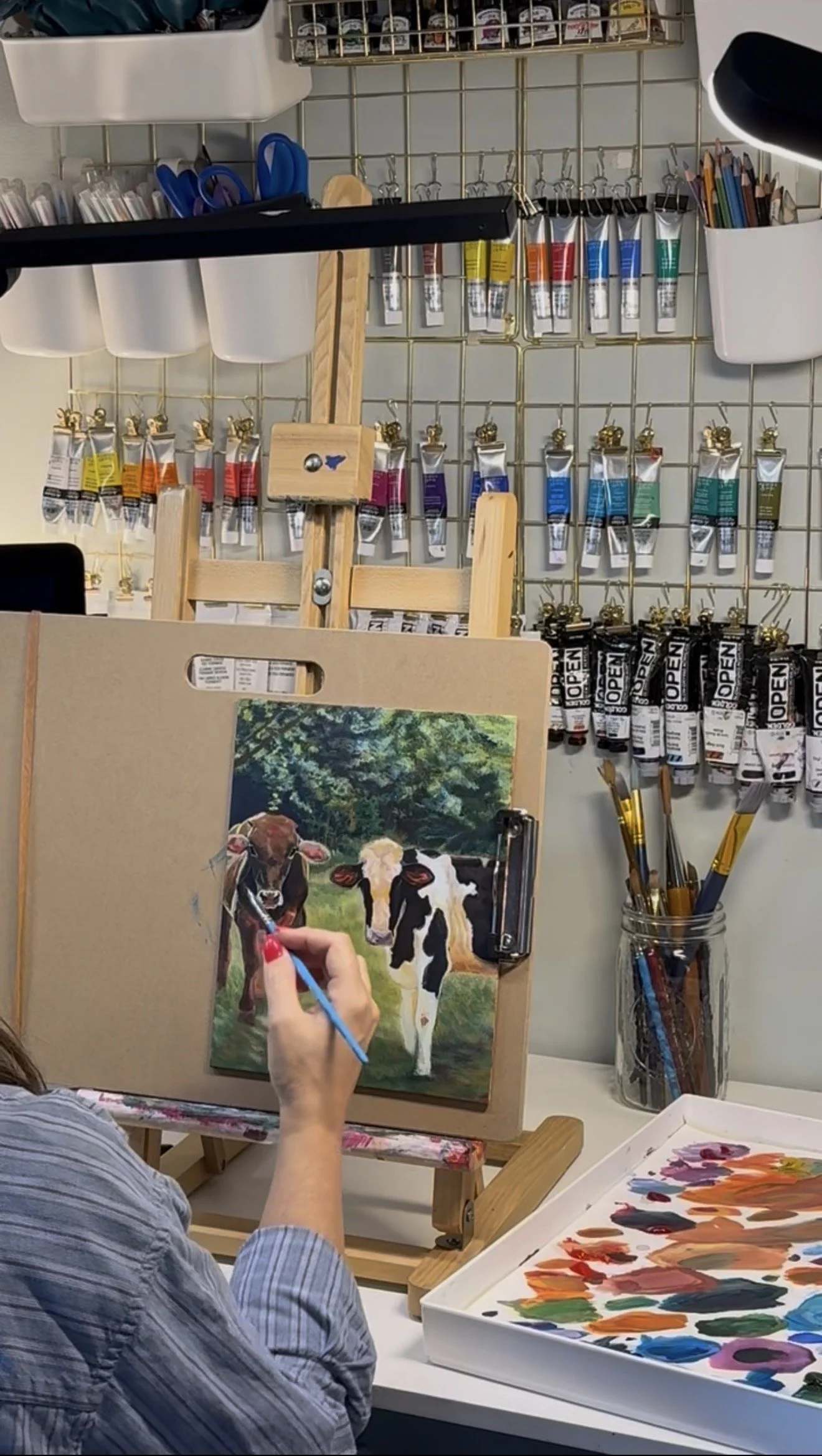 Person painting a realistic portrait of two cows on canvas, with art supplies and colorful paints visible on a table and shelves in the background.