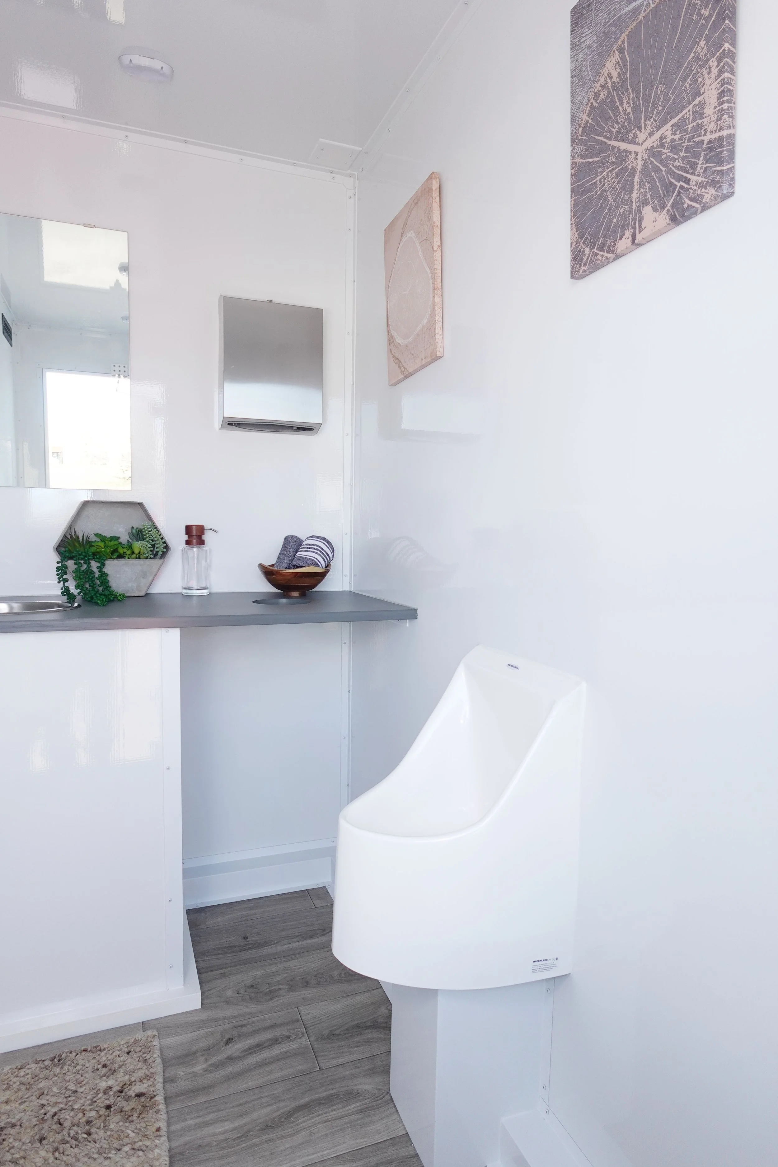 A modern, minimalist bathroom with white walls, a white toilet with a sleek design, a gray countertop, and a small bathroom vanity with a mirror. The wall features two pieces of abstract artwork, a hexagonal planters with greenery, a soap dispenser, and a bowl with towels. The flooring is wood-look gray tiles.