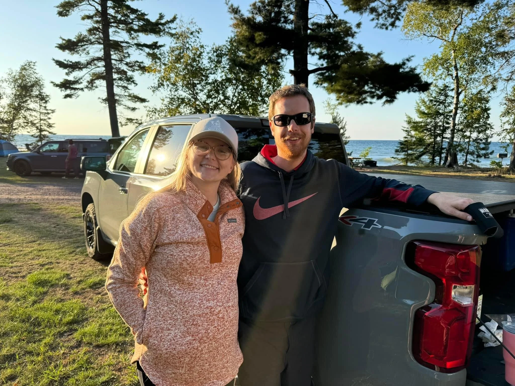 A smiling couple taking a selfie outdoors near a lake with cars parked behind them. The woman is wearing glasses, a light jacket with orange accents, and a beige cap. The man, wearing sunglasses and a black hoodie with red accents, is standing next to the woman. They are standing next to a gray pickup truck, with trees and the lake in the background during the late afternoon or early evening.