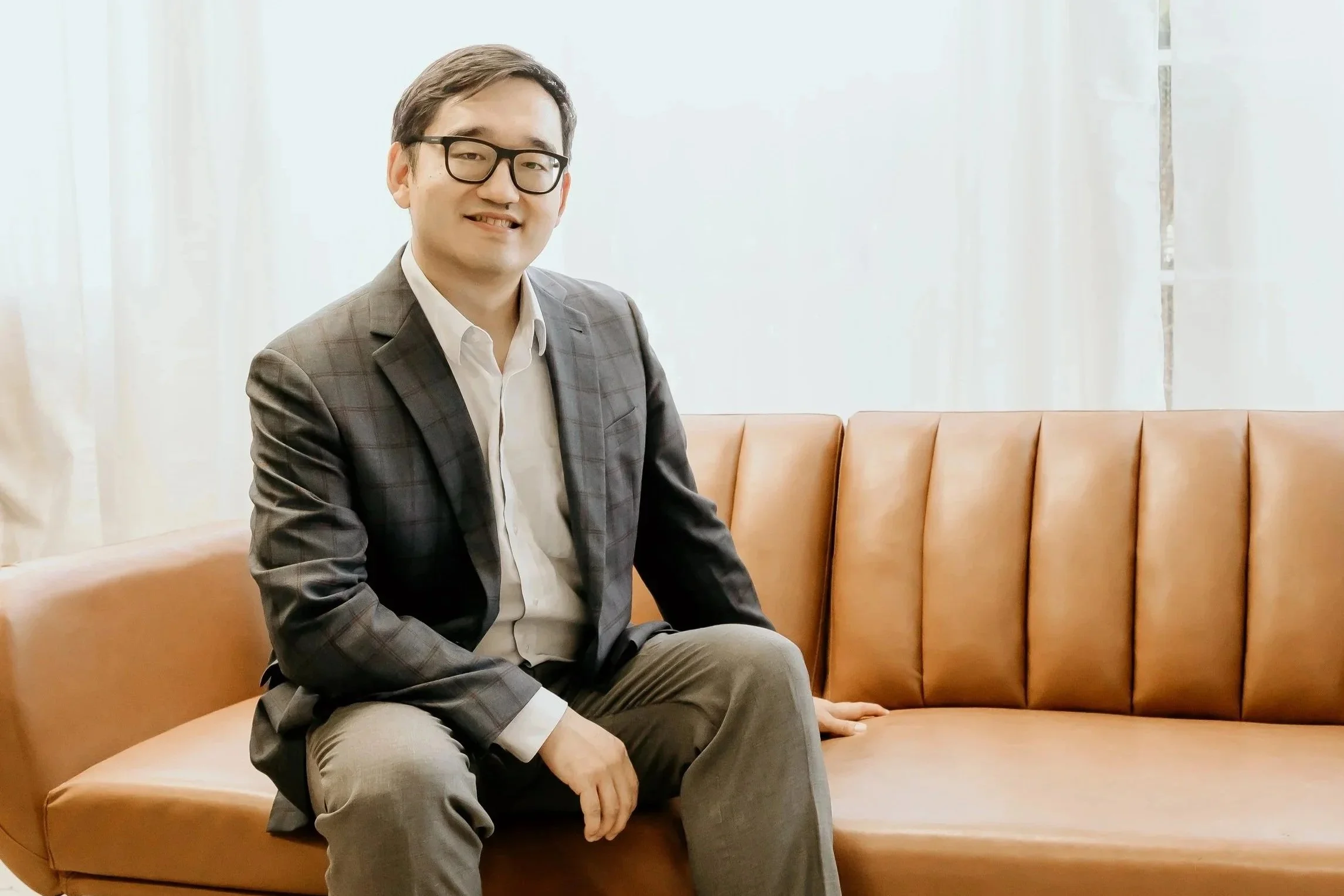 A man in a gray checkered suit, white shirt, and glasses sitting on a tan leather bench in a room with white curtains and a white wall, smiling at the camera.