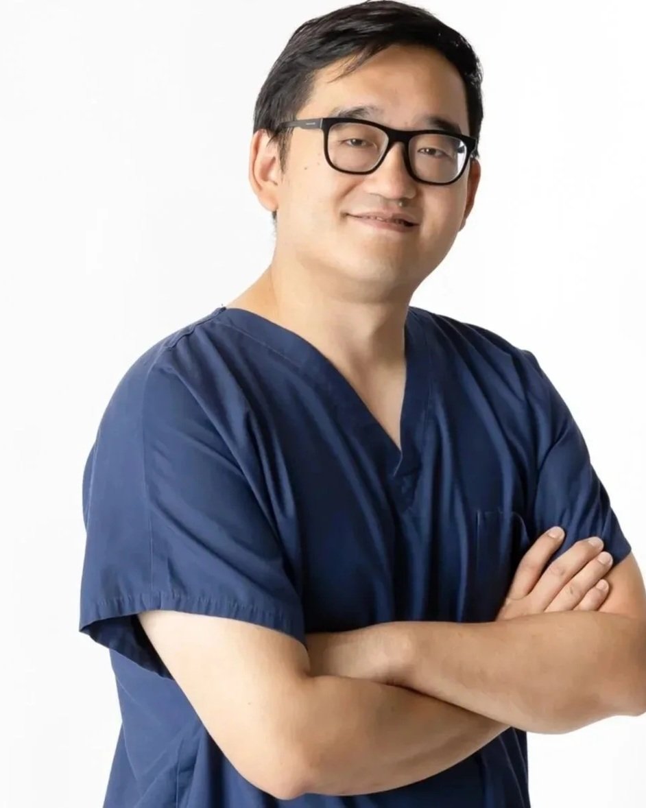 A portrait of a male healthcare professional in navy scrubs with crossed arms, wearing glasses, smiling, and standing against a white background.