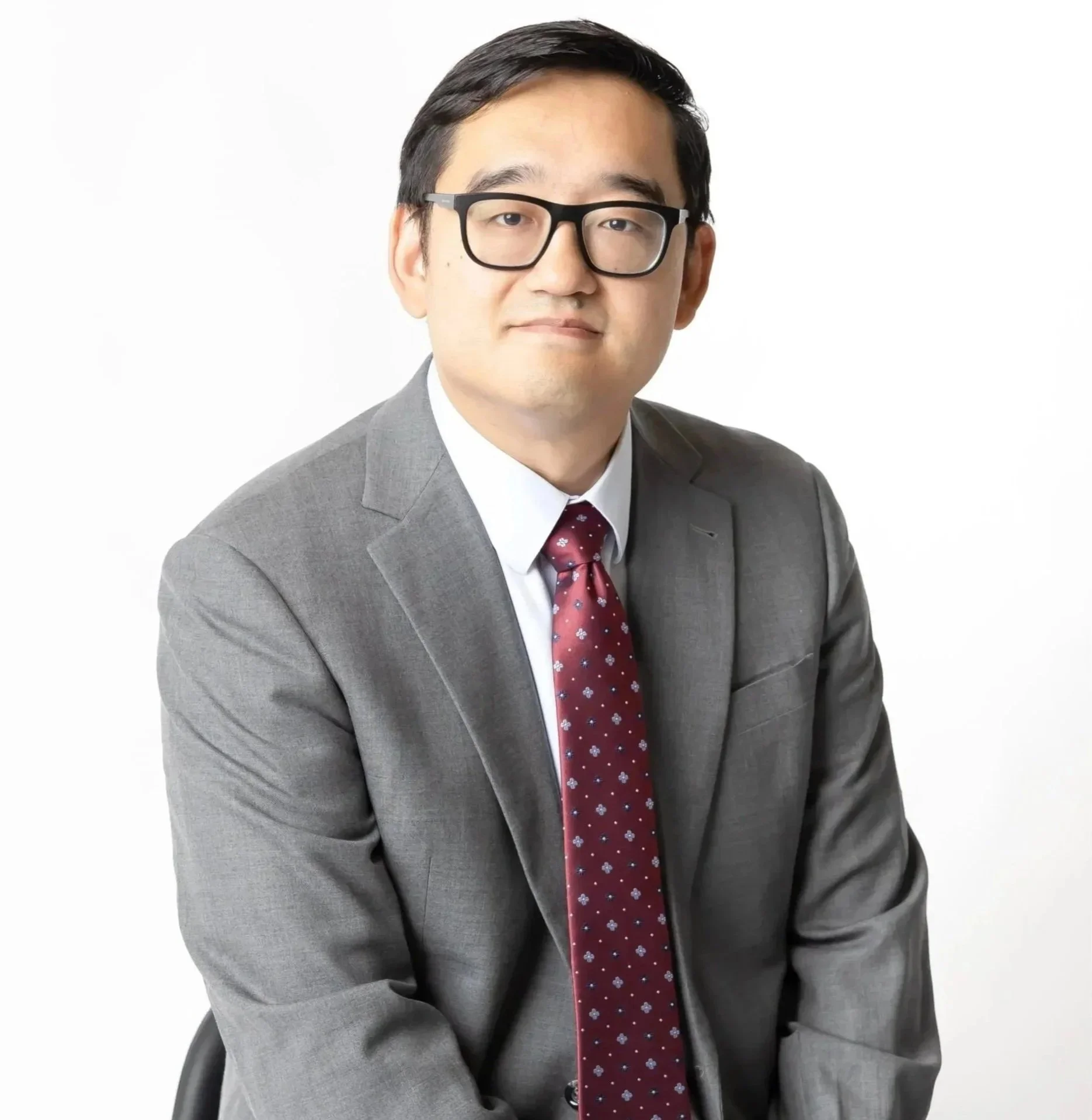 A man with black hair, glasses, wearing a gray suit, white shirt, and red tie with white and blue patterns, sitting in a black chair against a white background.