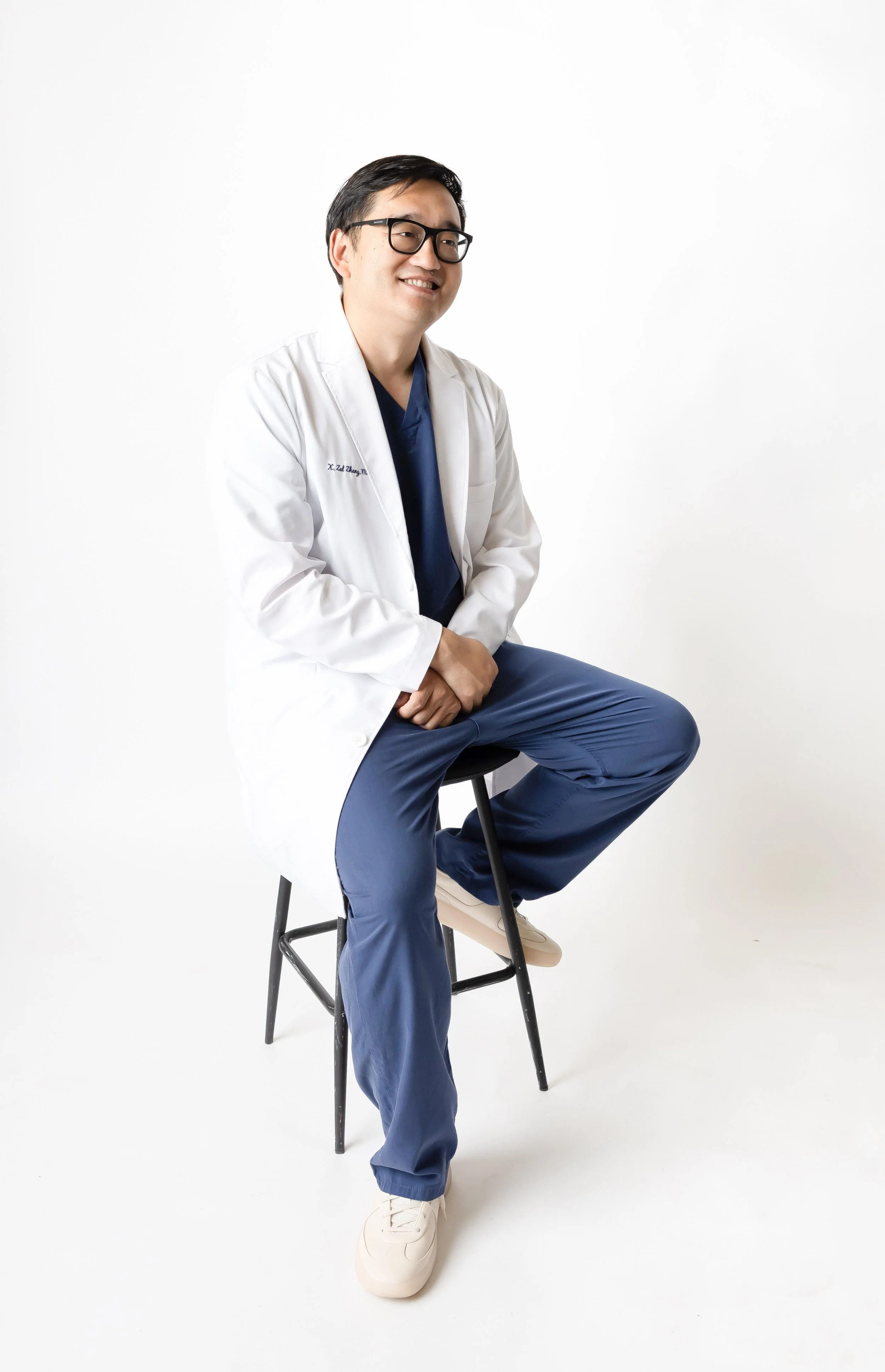 A male doctor wearing glasses, a white coat over blue scrubs, seated on a stool against a white background, smiling and looking to the side.