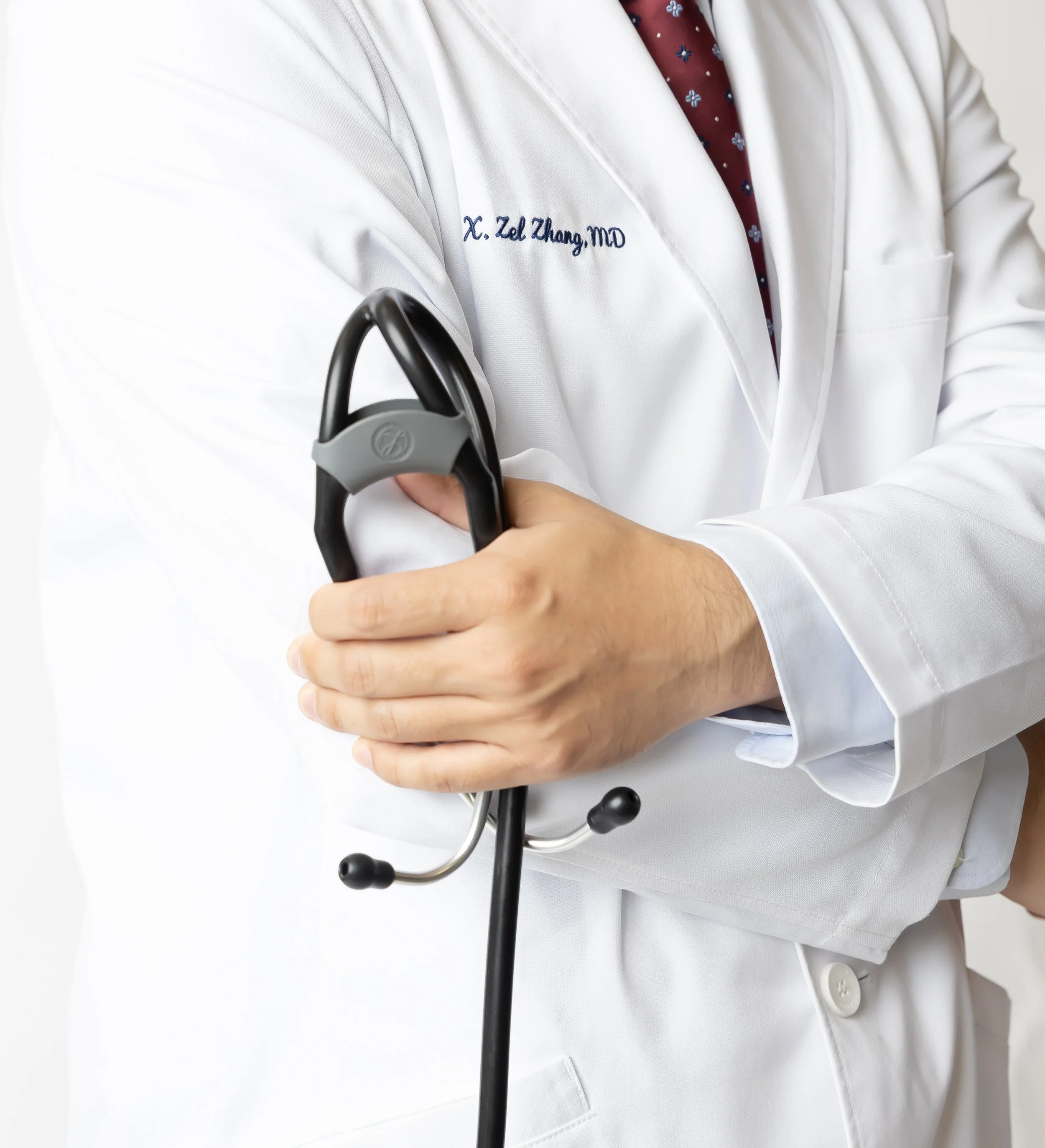 Close-up of a doctor wearing a white coat with embroidered name and title, holding a stethoscope, against a plain light background.