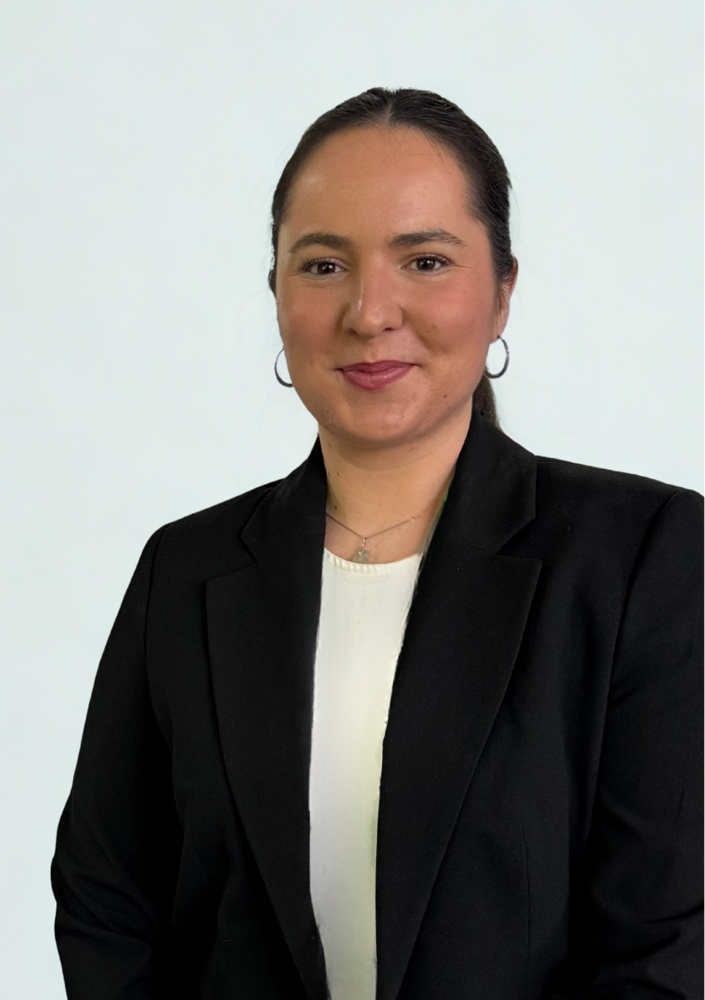 A woman with dark hair pulled back, wearing hoop earrings, a white top, and a black blazer, standing against a plain white background.