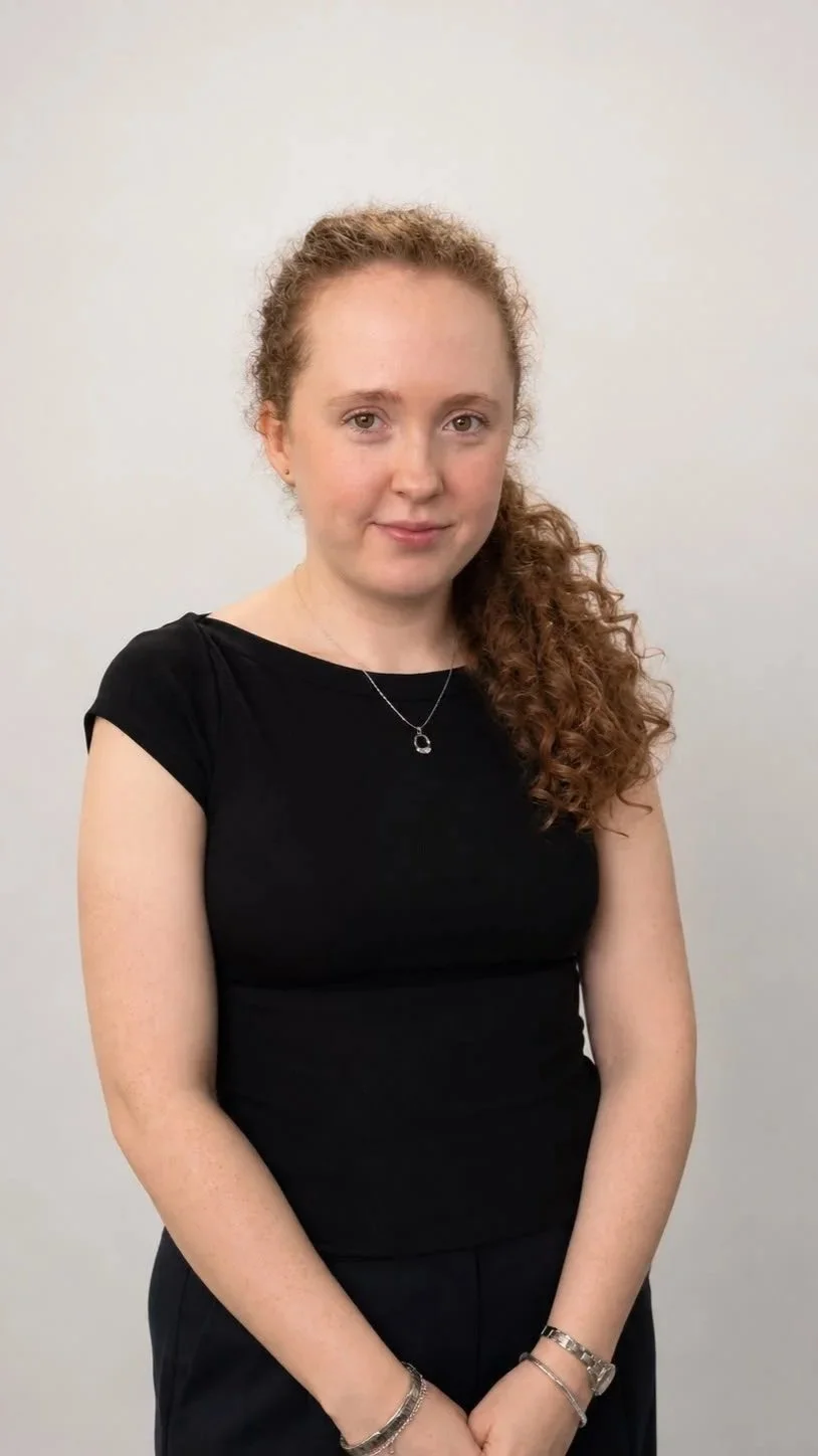 A woman with curly red hair wearing a black top and jewelry posing against a plain white background.