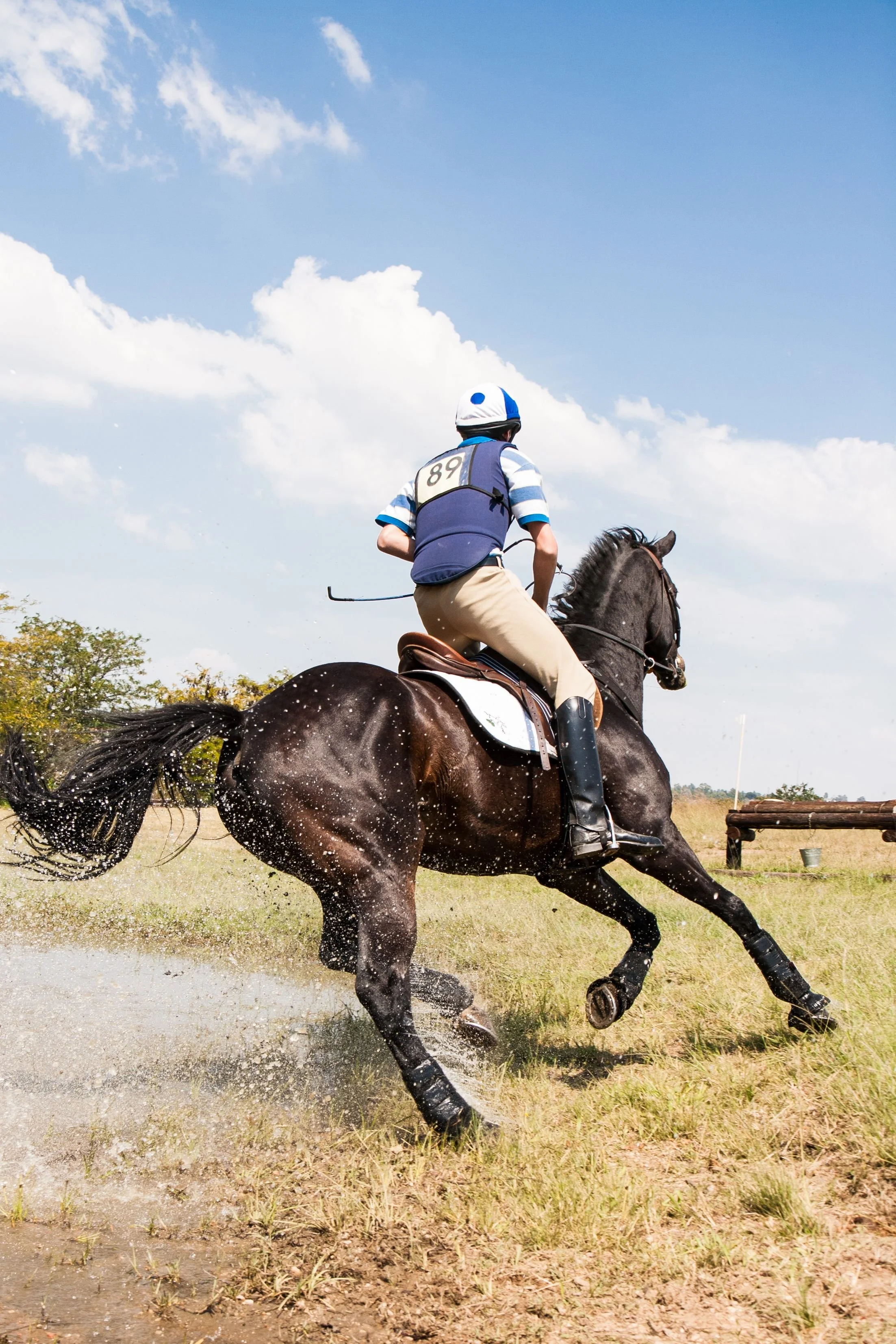 A person riding a horse at a gallop on a grassy field, with water splashing behind the horse's legs and a partly cloudy sky overhead.