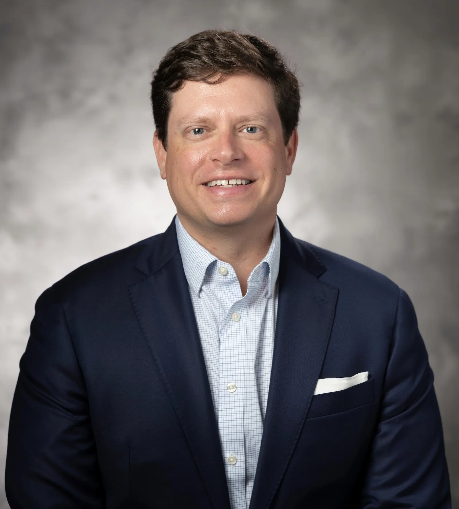 Headshot of a smiling man with dark hair, dressed in a navy blazer, white dress shirt, and pocket square, against a gray background.