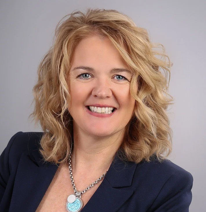 A smiling woman with blonde, curly hair, wearing a jewelled necklace and a navy blazer against a plain gray background.