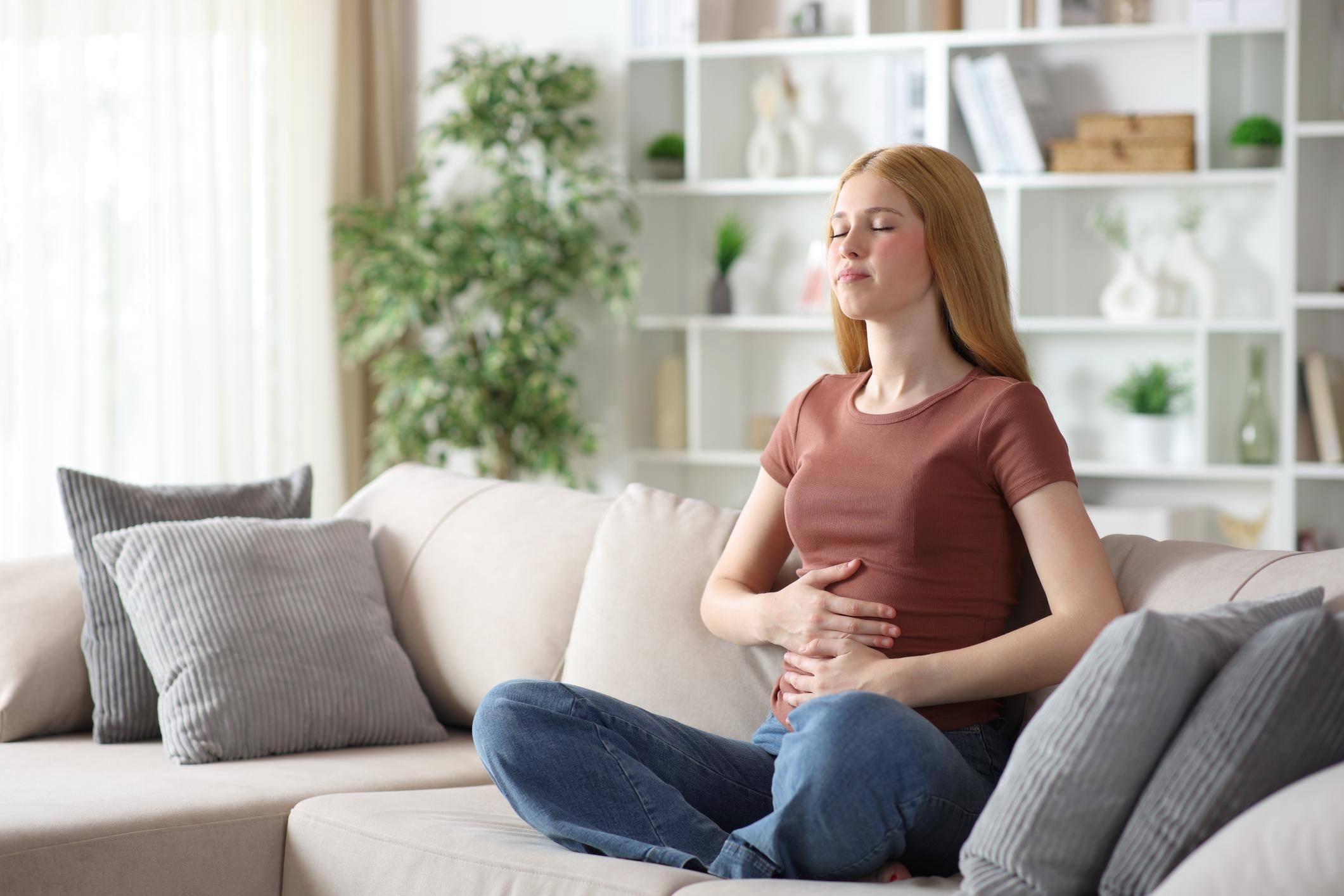 A woman sitting on a beige sofa with her eyes closed and hands on her stomach, possibly experiencing stomach discomfort, in a bright living room with white bookshelves and green plants.