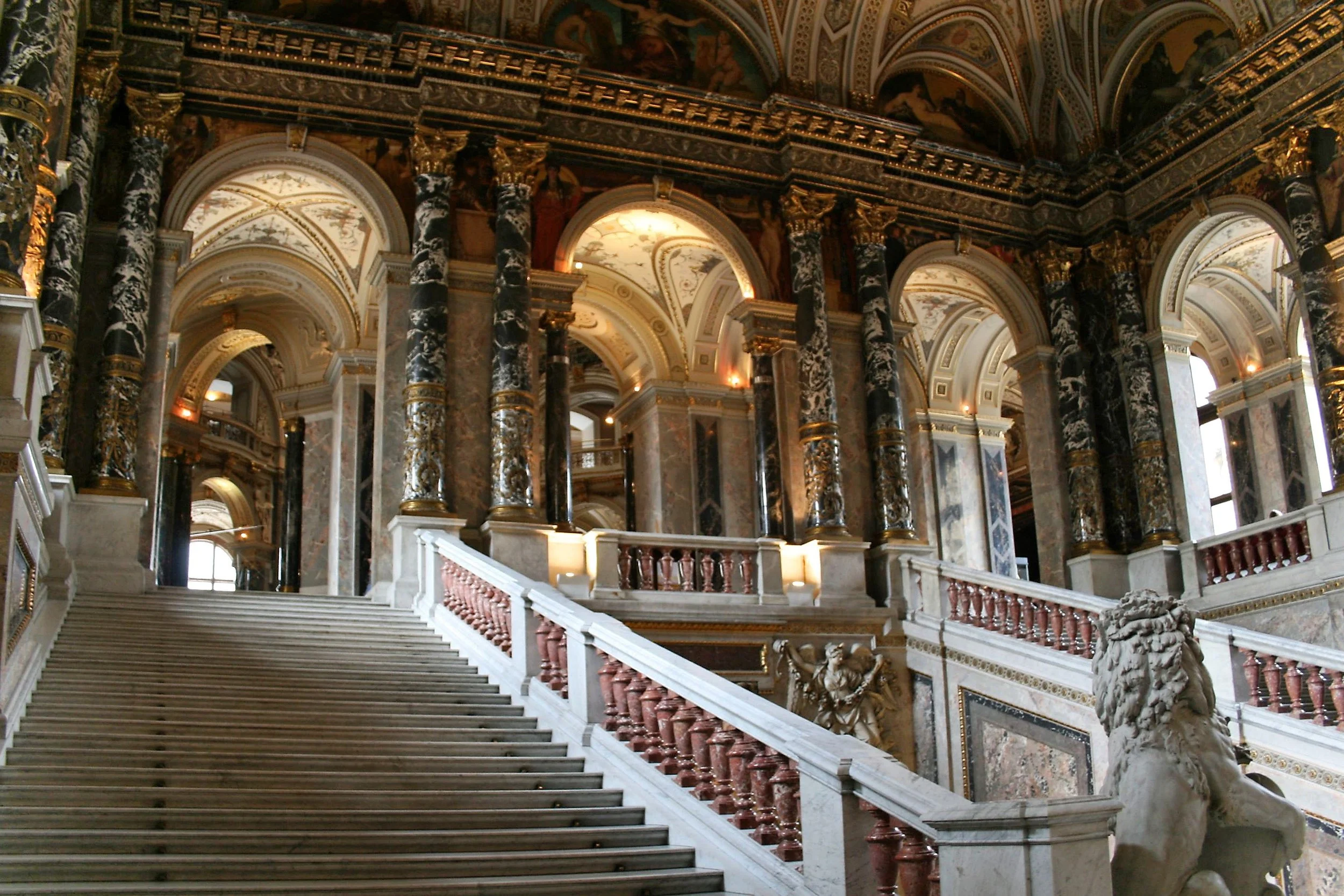 Elegant grand staircase inside a baroque-style palace or cathedral, with marble and gilded ornamentation, large columns, and detailed murals on the ceiling.