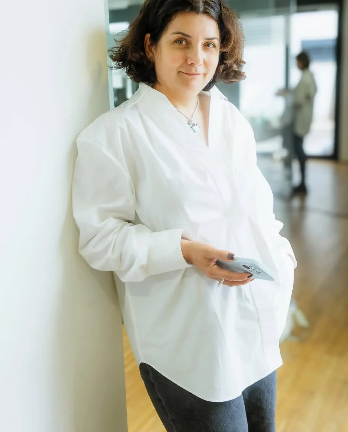 A woman with short, dark, curly hair, wearing a white blouse and gray jeans, leaning against a wall in an indoor setting, holding a smartphone in her hand, with a blurred background of another person in a white coat.