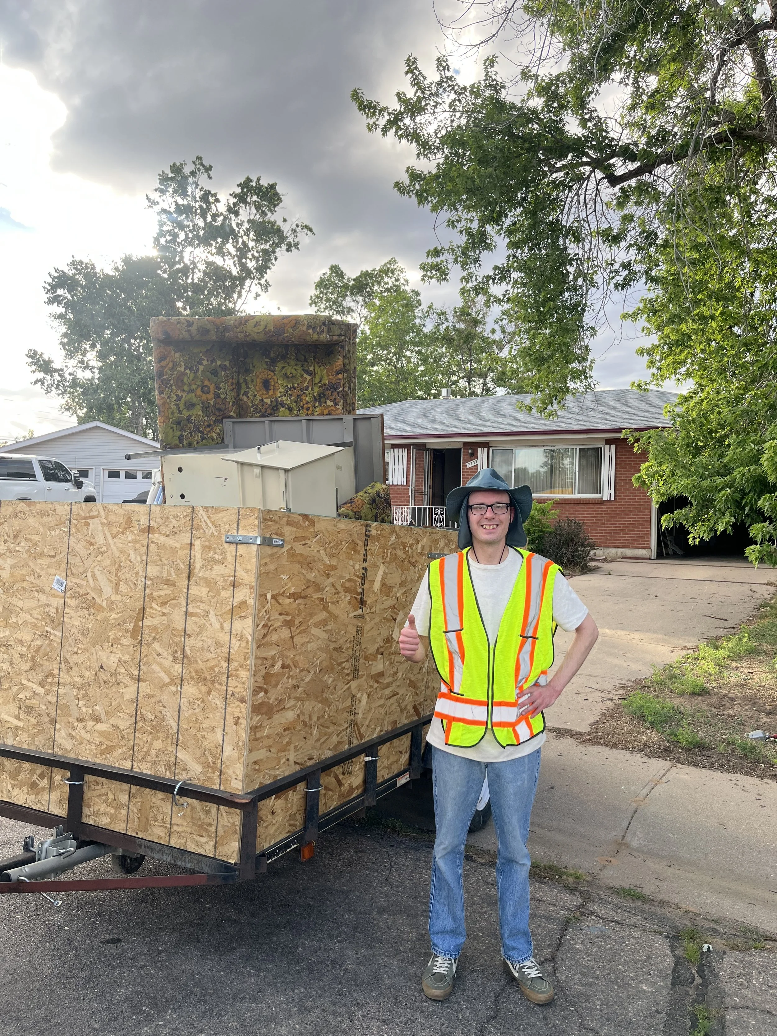 A man in a hard hat and safety vest giving a thumbs-up pose beside a trailer loaded with furniture, including a floral armchair and other household items, parked on a residential street.