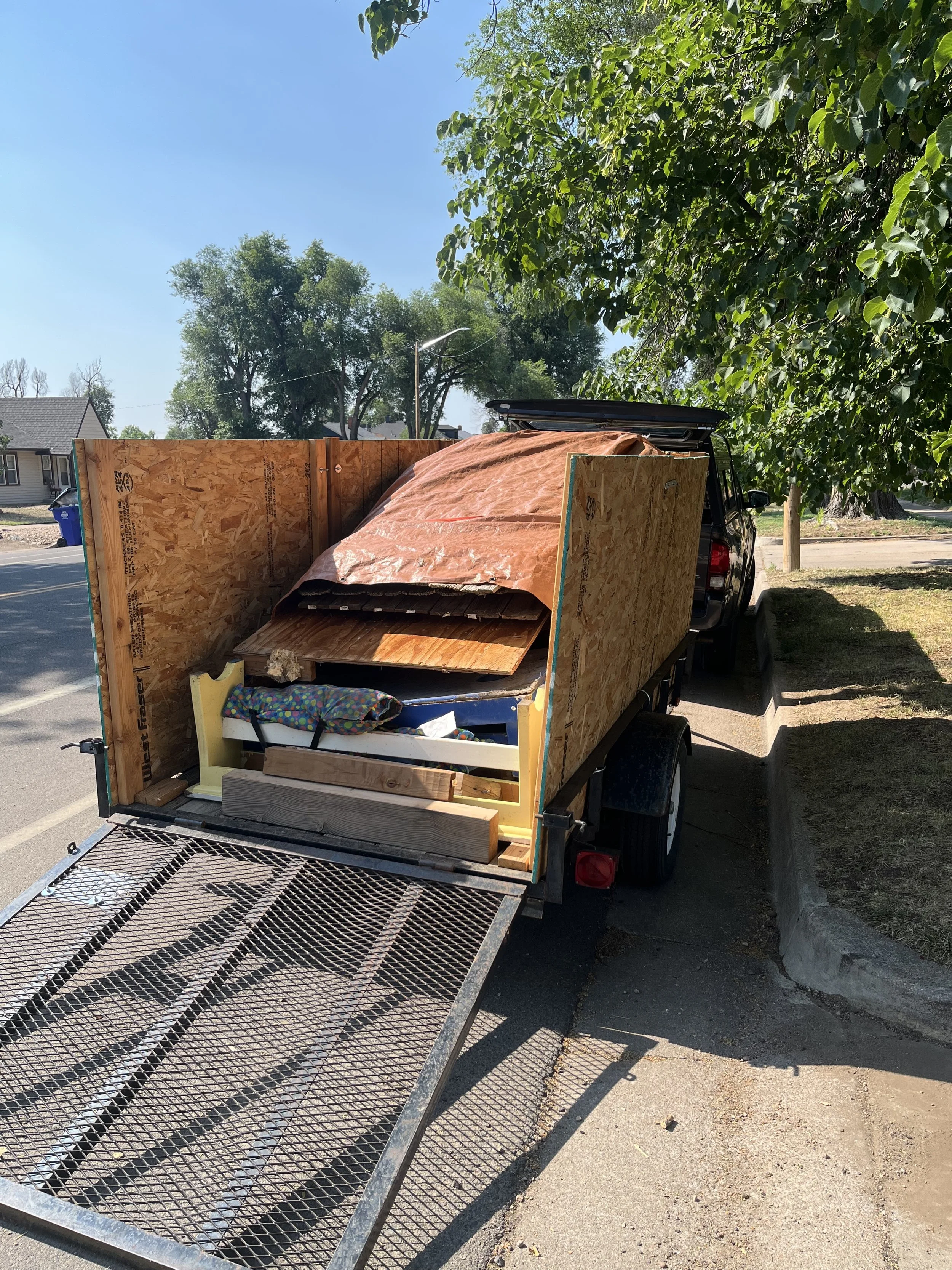 A trailer loaded with wooden and construction materials parked on the side of a street, with trees and houses in the background under a clear blue sky.