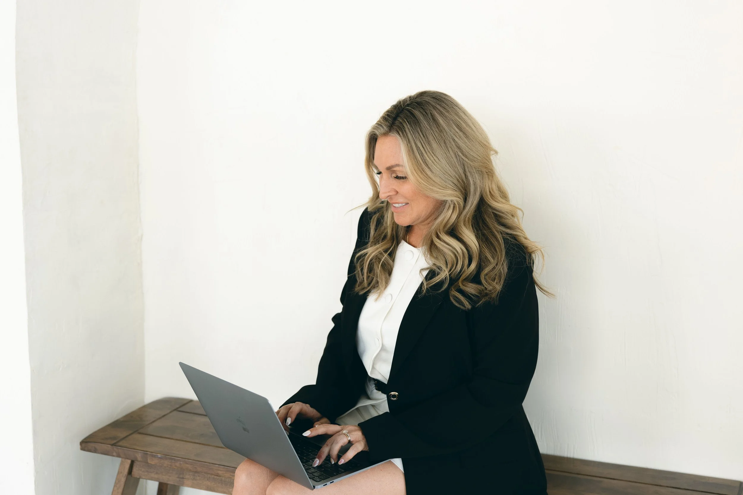 A woman with long wavy blonde hair working on a silver laptop while seated on a wooden bench, wearing a black blazer and white blouse, against a plain white wall.