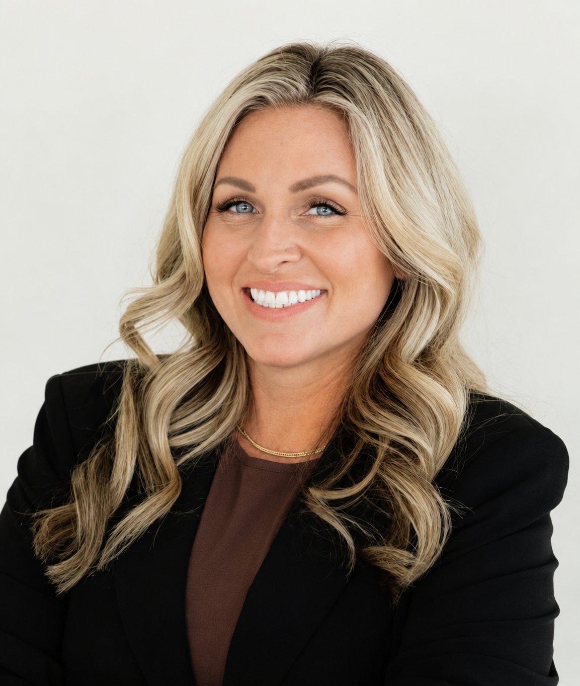 Professional woman with blonde, wavy hair smiling, wearing a black blazer and gold necklace against a plain white background.