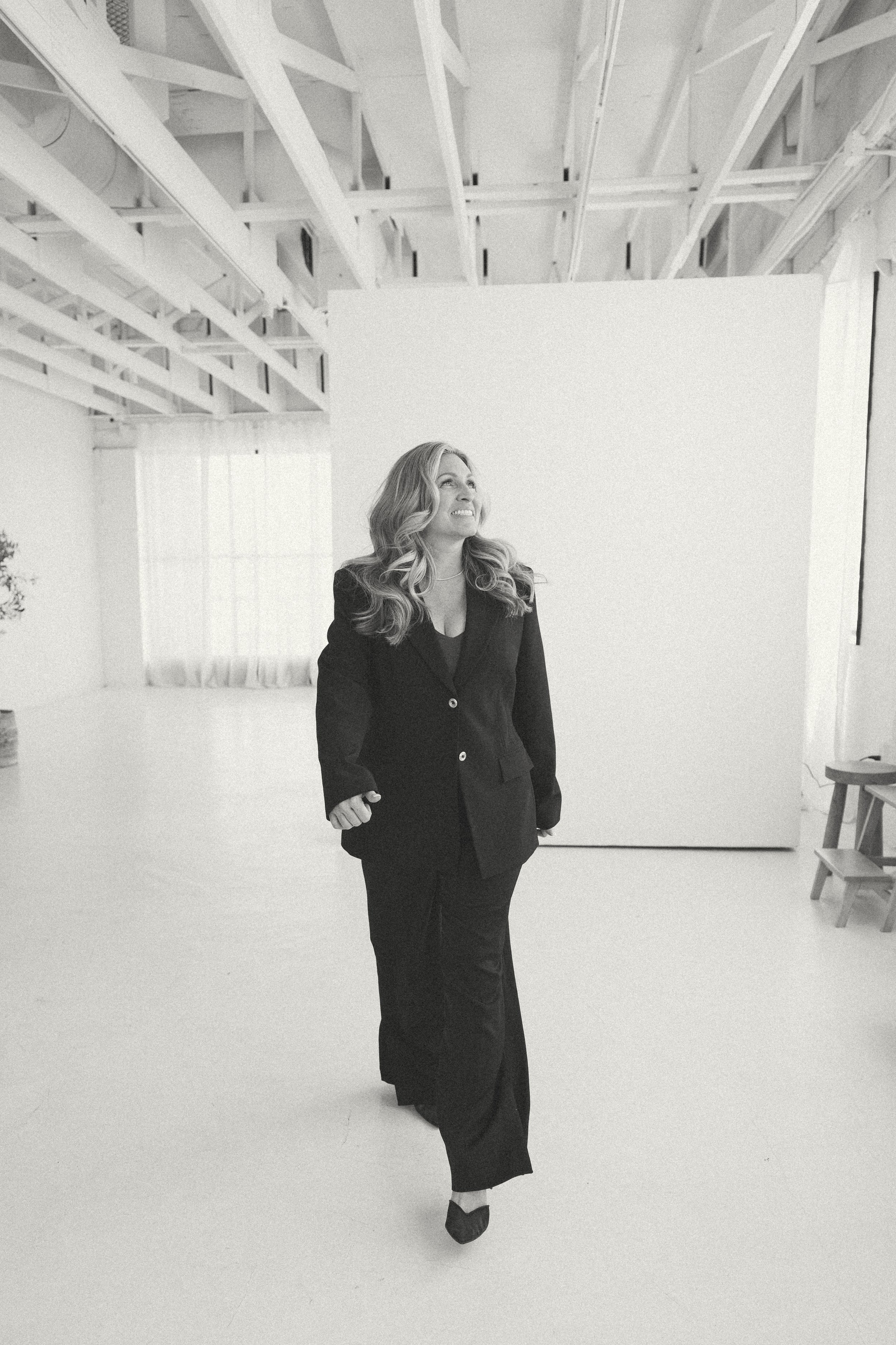 A woman with long, wavy hair wearing a dark professional suit, standing in a spacious, airy room with a high ceiling with exposed white beams, a large white wall, and a window with sheer curtains, smiling and looking slightly upwards.