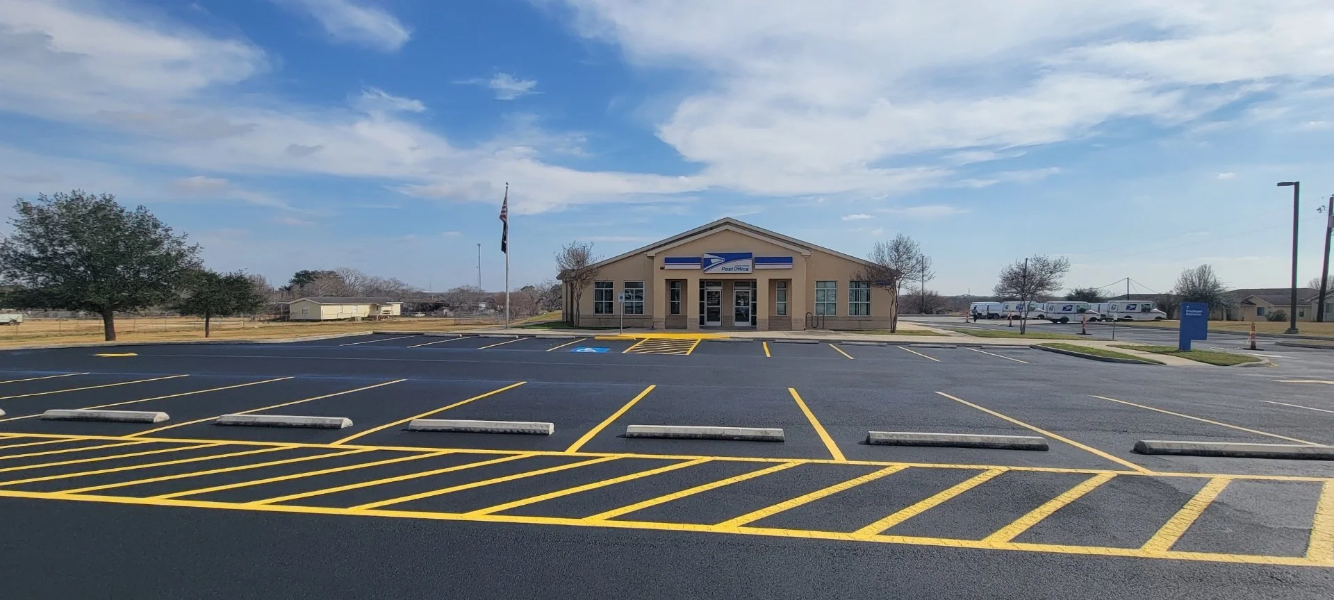 Empty parking lot with yellow-striped parking spaces, a small post office building with a sign, an American flag, and USPS logo, surrounded by clear sky and a few trees.