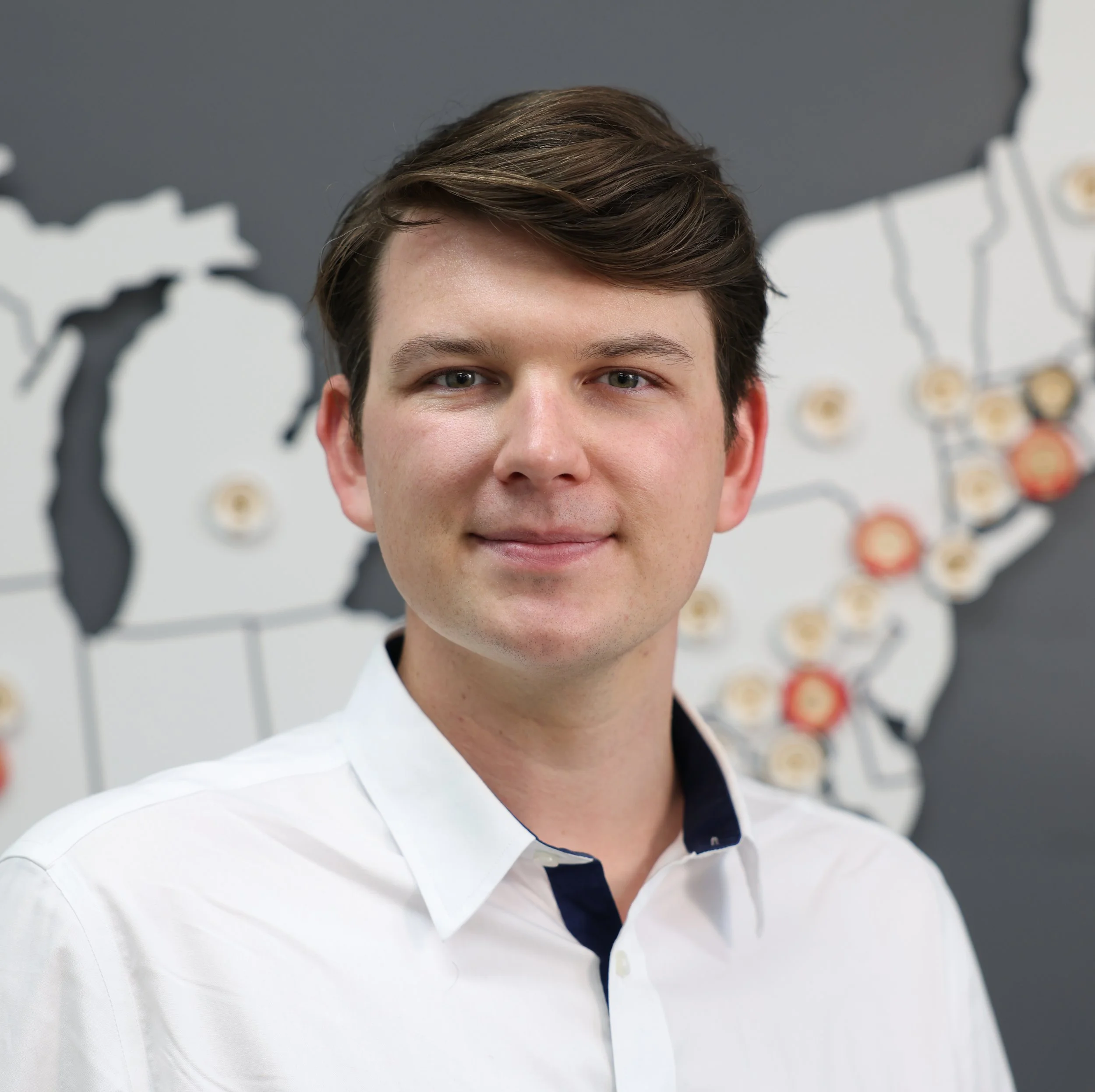 Headshot photograph of Alex Bruens, taken in 2026. Alex is looking directly at camera, and has brown hair and a soft smile. Alex is wearing a white shirt and stands in front of a map.