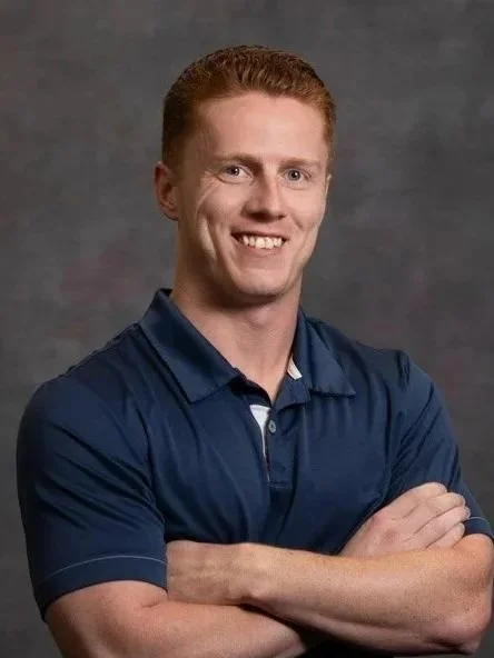 Smiling man with red hair and fair skin wearing a navy blue polo shirt, standing with arms crossed against a gray background.