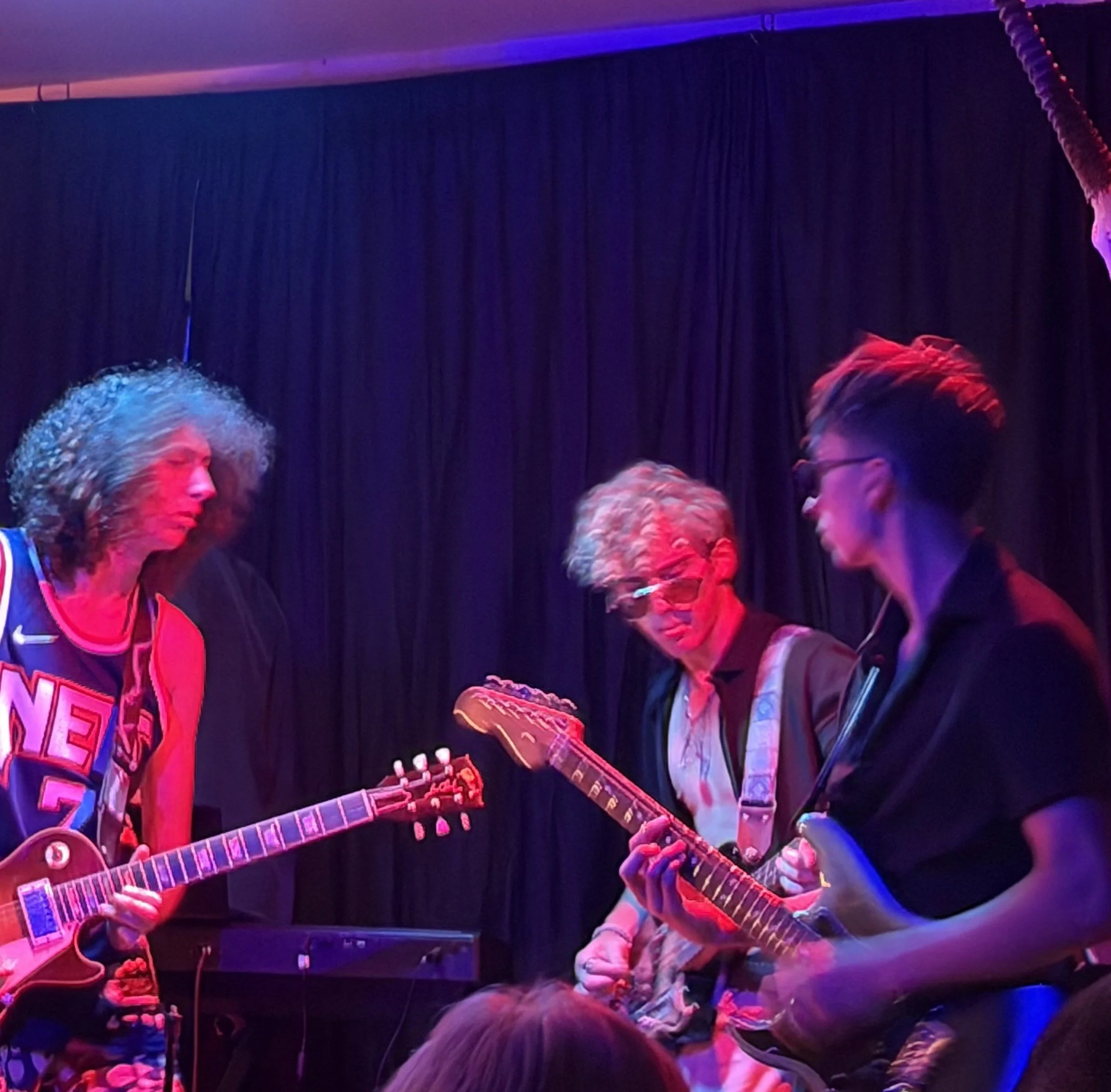 People playing electric guitars on a stage with dark curtains and colorful lighting.