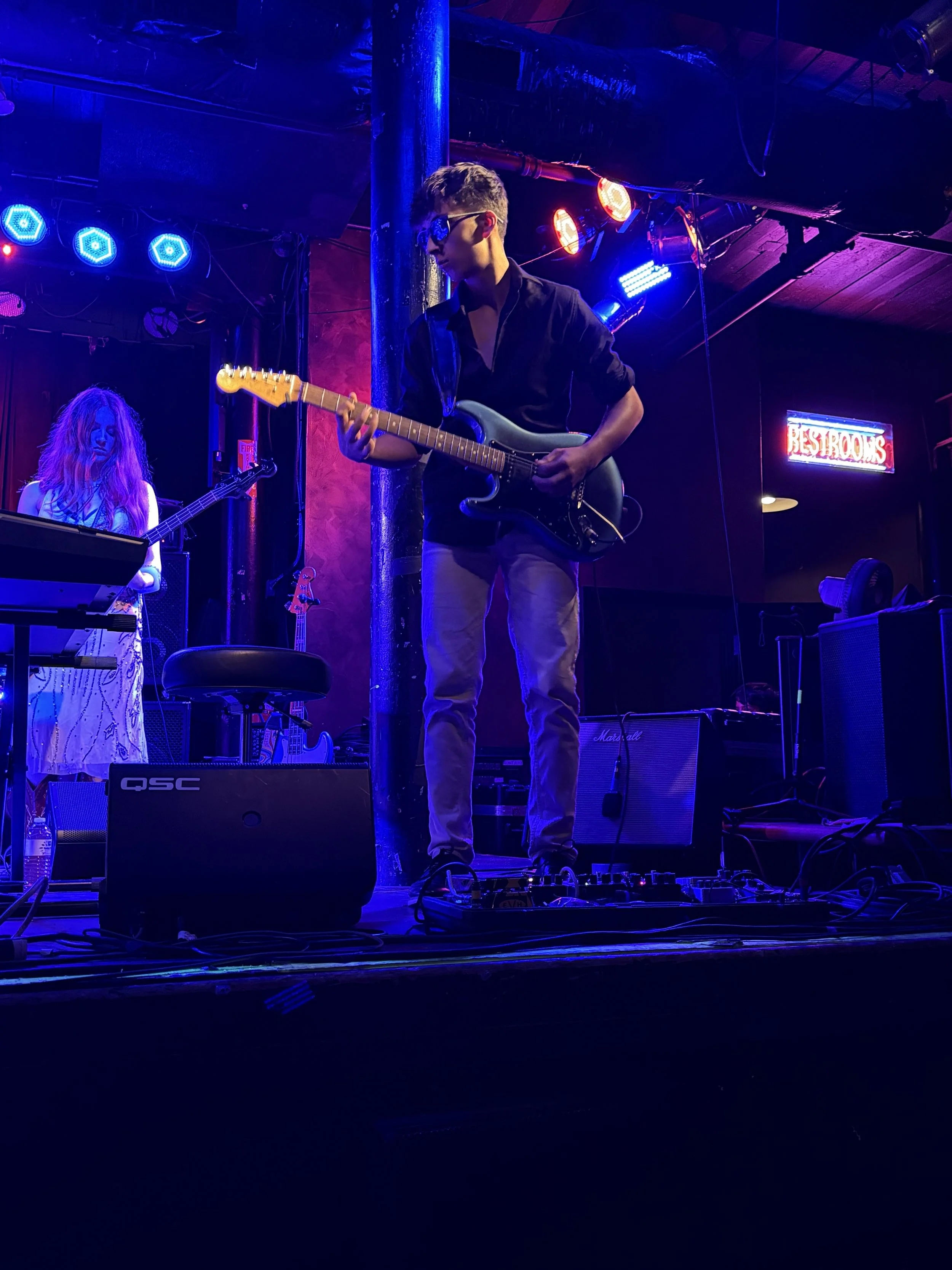 A young man wearing sunglasses playing an electric guitar on stage, with a woman in the background playing a bass guitar, and blue and red stage lights illuminating the scene.