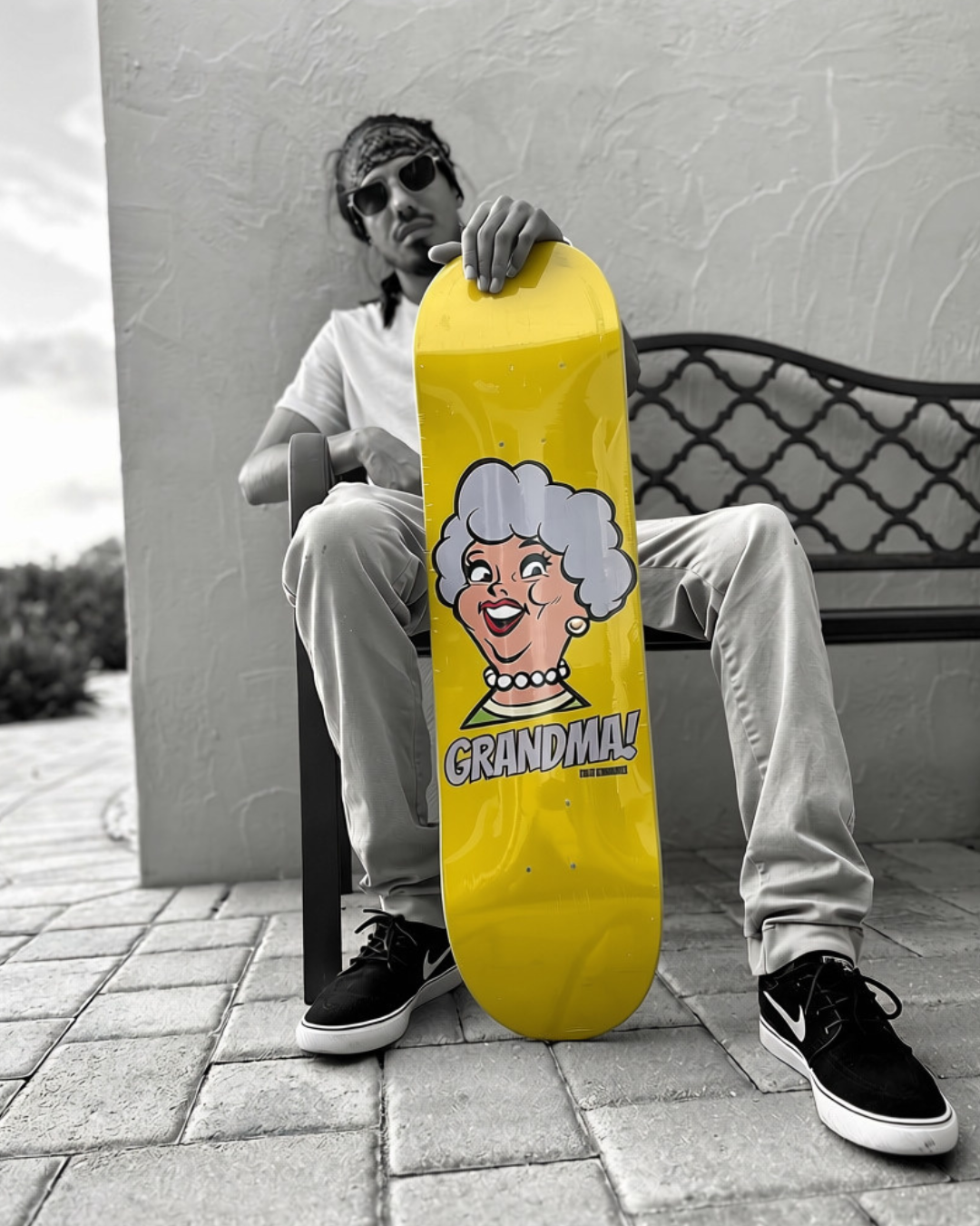 A person sitting on a bench holding a bright yellow skateboarding deck featuring a cartoon of an elderly woman's face with gray hair and the word "GRANDMA!" in bold letters. The photo is in black and white except for the skateboard.