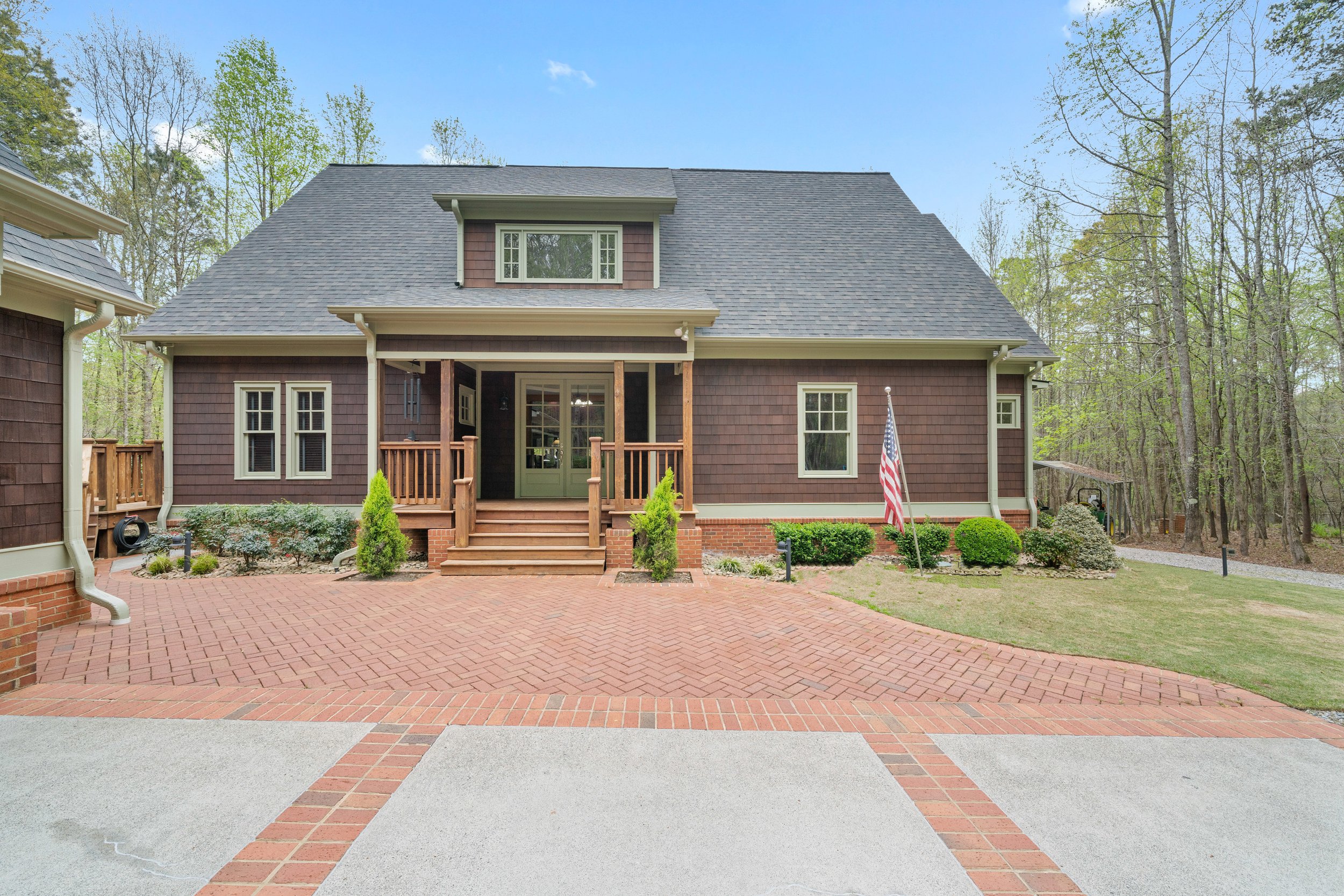 A two-story house with a gray roof, brown siding, and a small front porch with wooden steps. There are trees in the background, a brick and concrete driveway in the foreground, and an American flag on a pole in the yard.