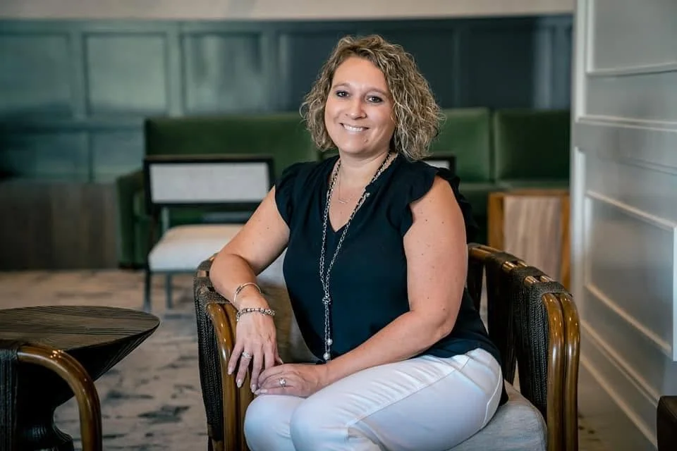A woman with curly blonde hair sitting on a chair in a modern indoor space, wearing a black top, white pants, and jewelry, smiling at the camera.