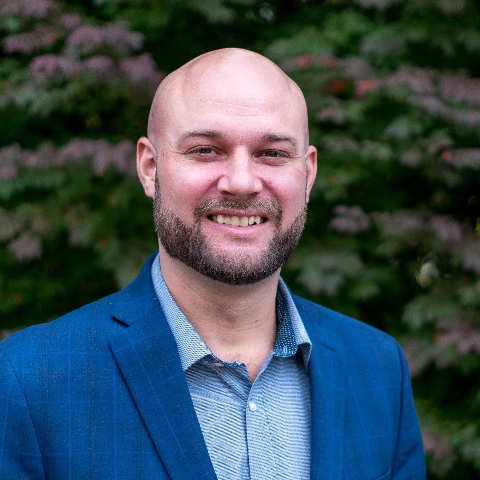 A smiling man in a blue plaid suit and light blue shirt standing outdoors with blurred greenery and flowers in the background.