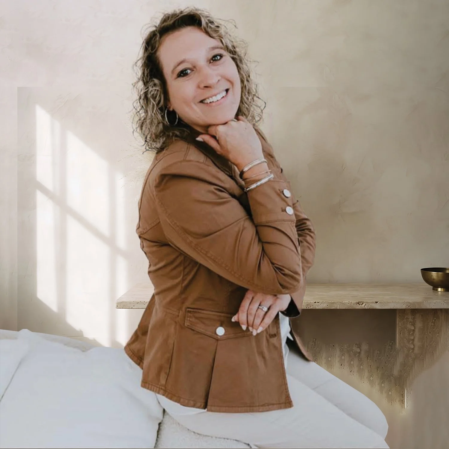 A woman with curly hair smiling, wearing a brown jacket, standing indoors near a beige wall with a marble shelf and a small bowl.