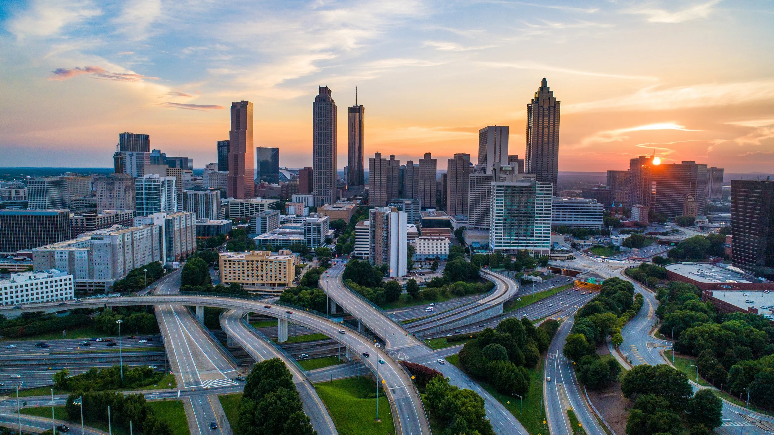 A city skyline during sunset with high-rise buildings, an intricate network of roads and highways, and greenery in the foreground.