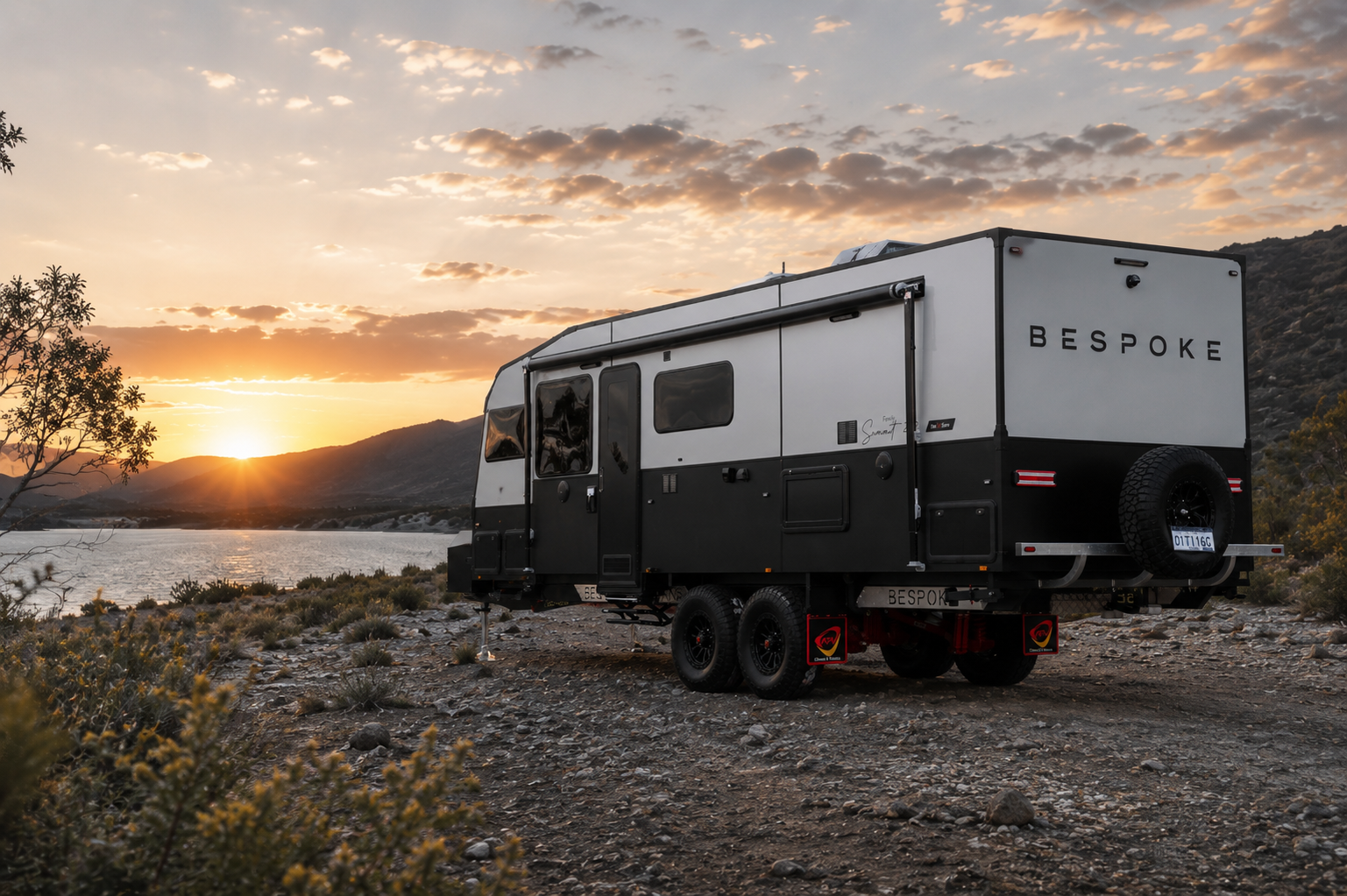 A black and white Bespoke Caravans 23 foot Family Summit model caravan parked on Australian rocky terrain near a body of water during sunset, with mountains and a partly cloudy sky in the background.