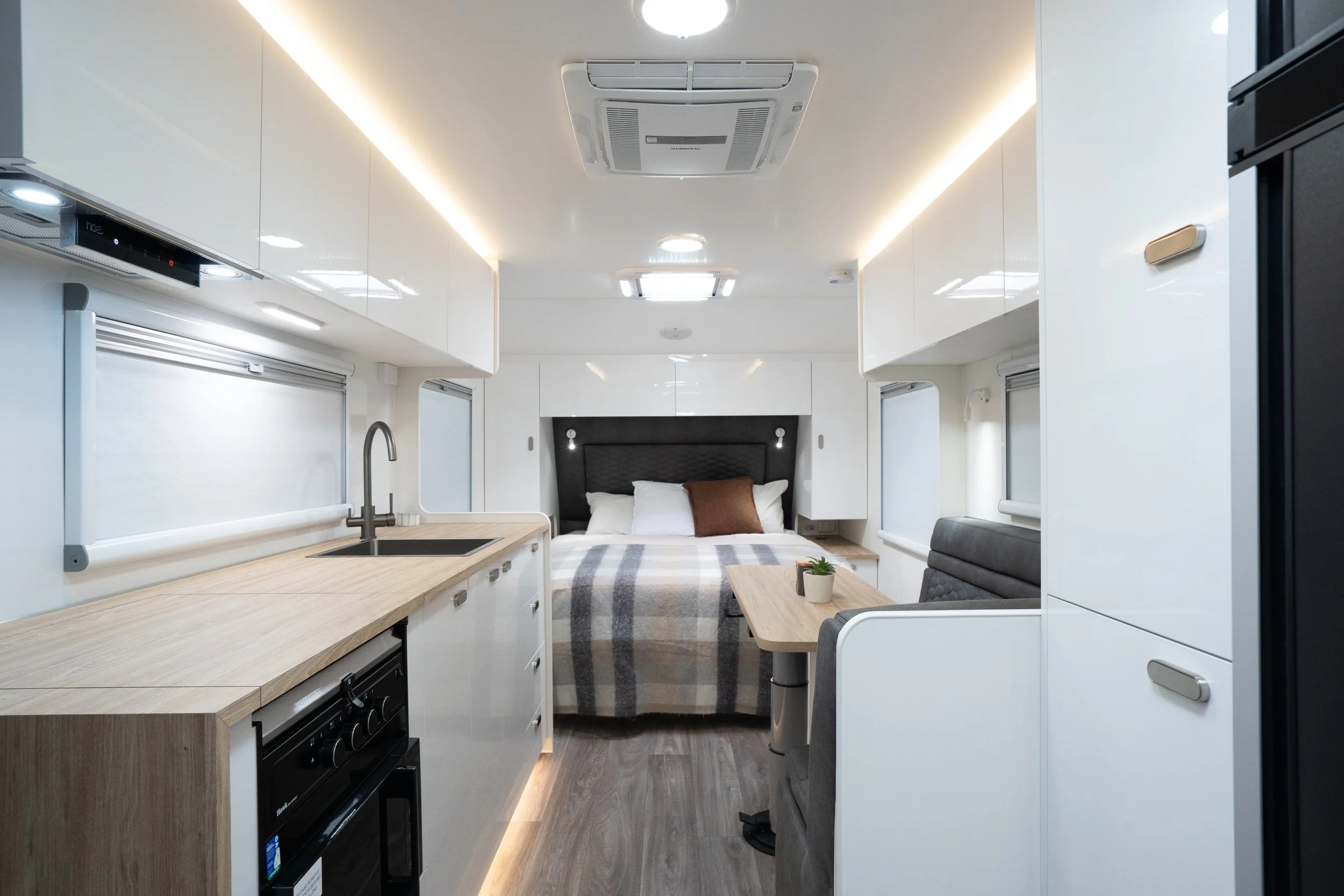North half of the interior of a modern Bespoke Caravans layout with a kitchen area, bed, dining space and sleek white cabinetry.