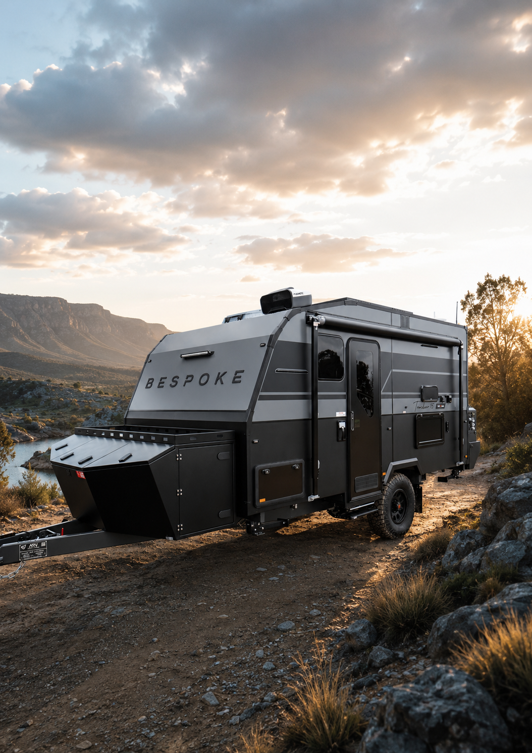 Bespoke Caravans Evoke model off-grid caravan parked on a dirt trail in a scenic outdoor setting with mountains, trees, and a river, during sunset.